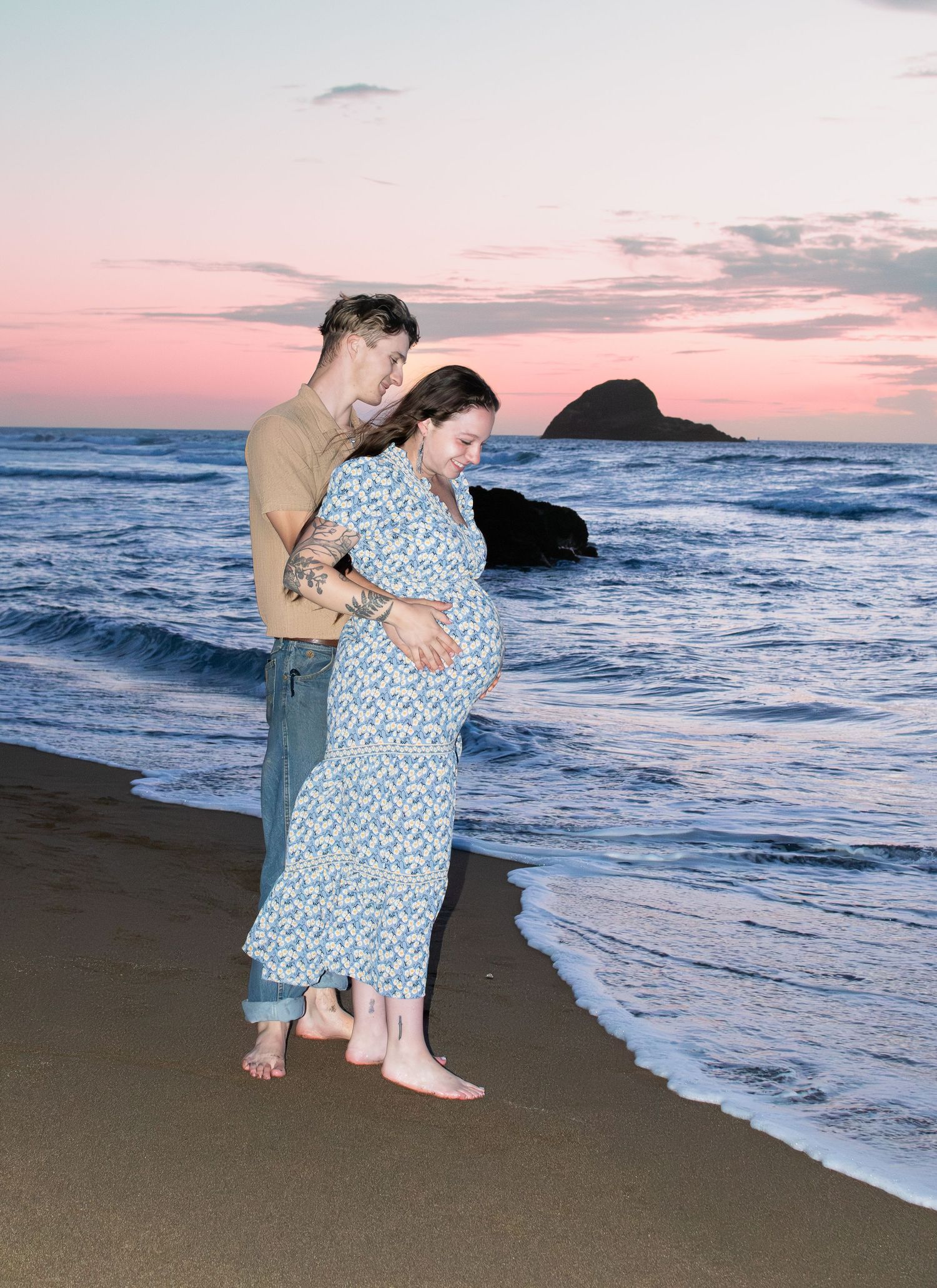 Pregnant couple standing barefoot on beach at sunset, man embracing woman in floral maxi dress near ocean waves.