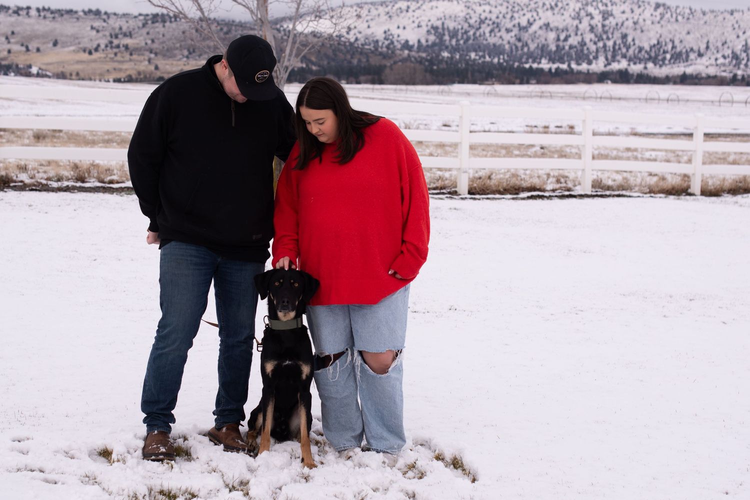 Couple in winter clothes standing with a dog in snowy field with white fence and mountains in background.