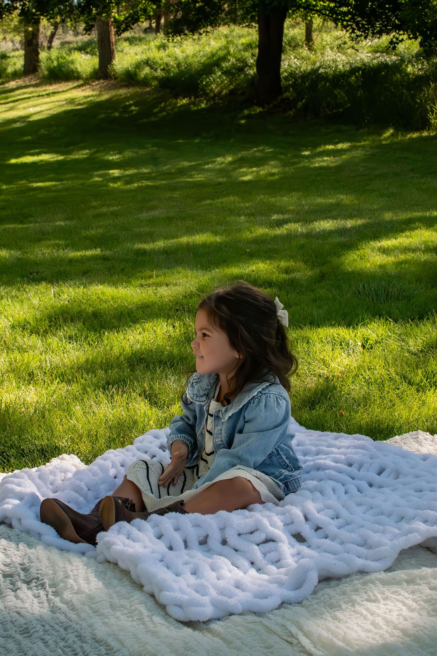 Young girl in denim jacket sits on chunky knit white blanket in sunny green park.