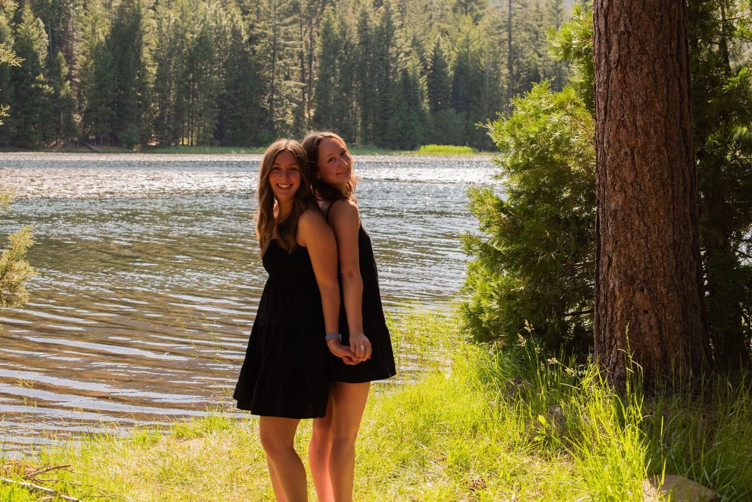 Two smiling women in black dresses stand back-to-back holding hands by a beautiful forest lake.