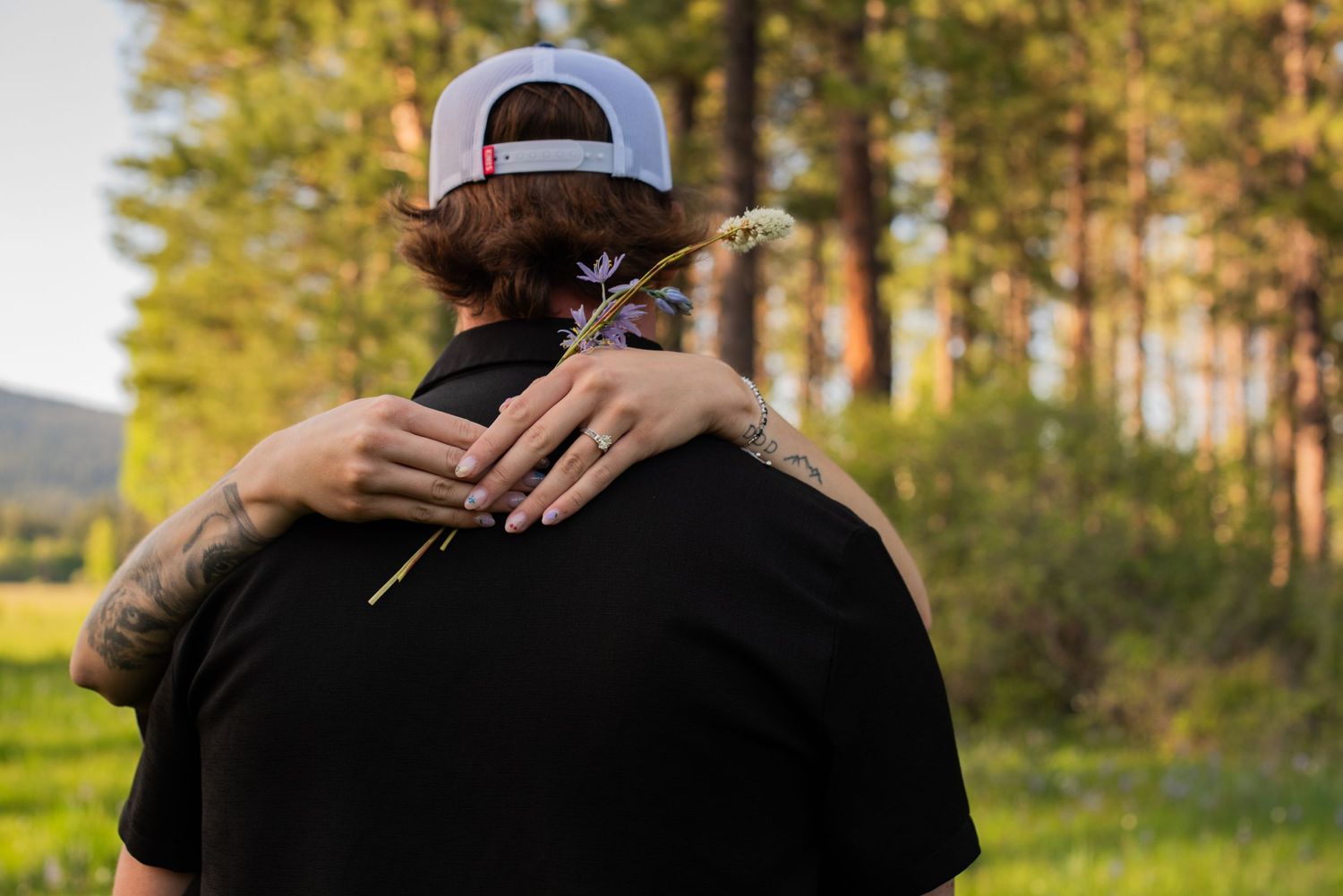 Close-up of a woman hugging a tattooed man holding a wildflower in a green outdoor setting.