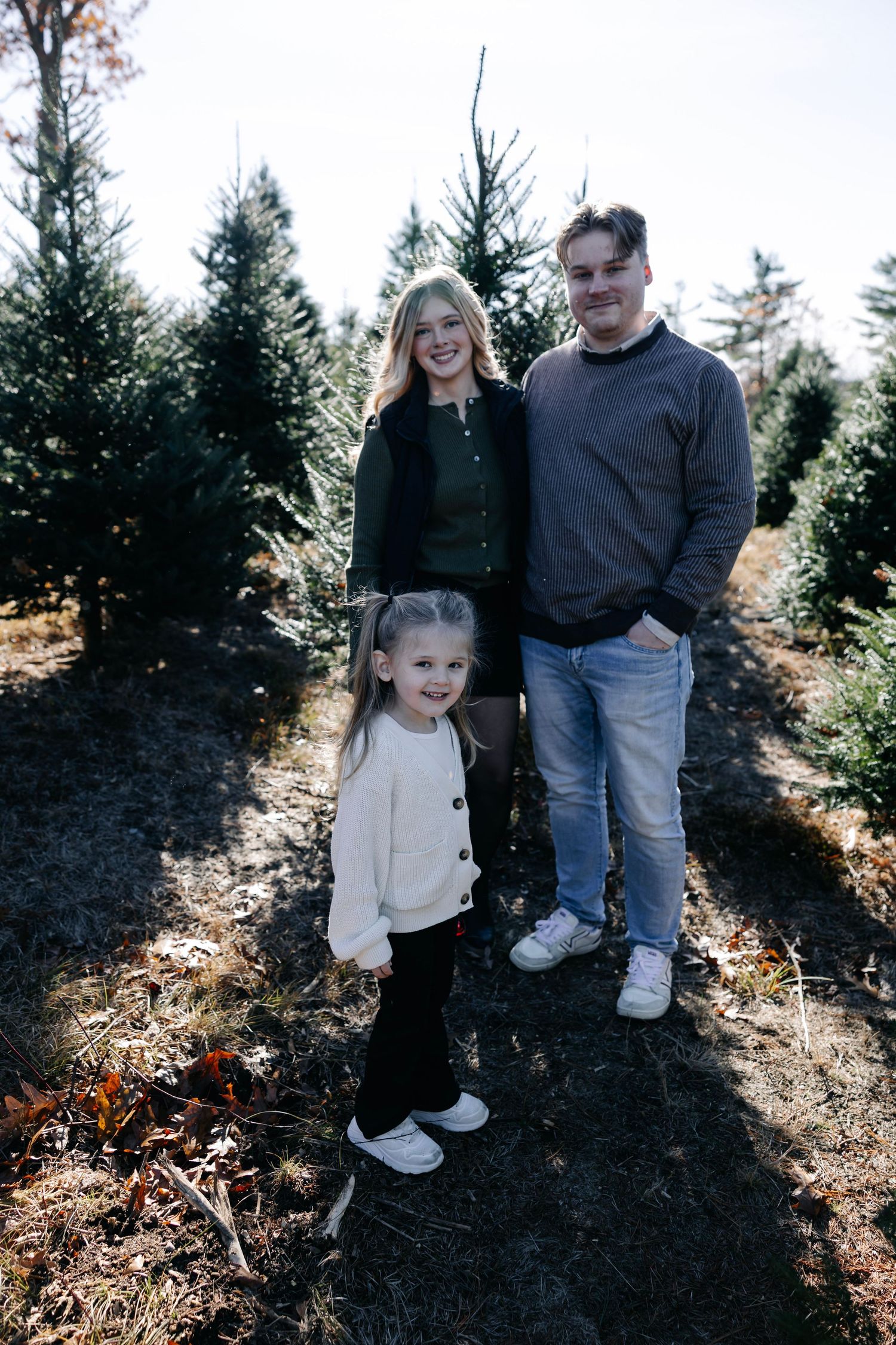 Family standing together on dark soil surrounded by evergreen trees during outdoor photoshoot.