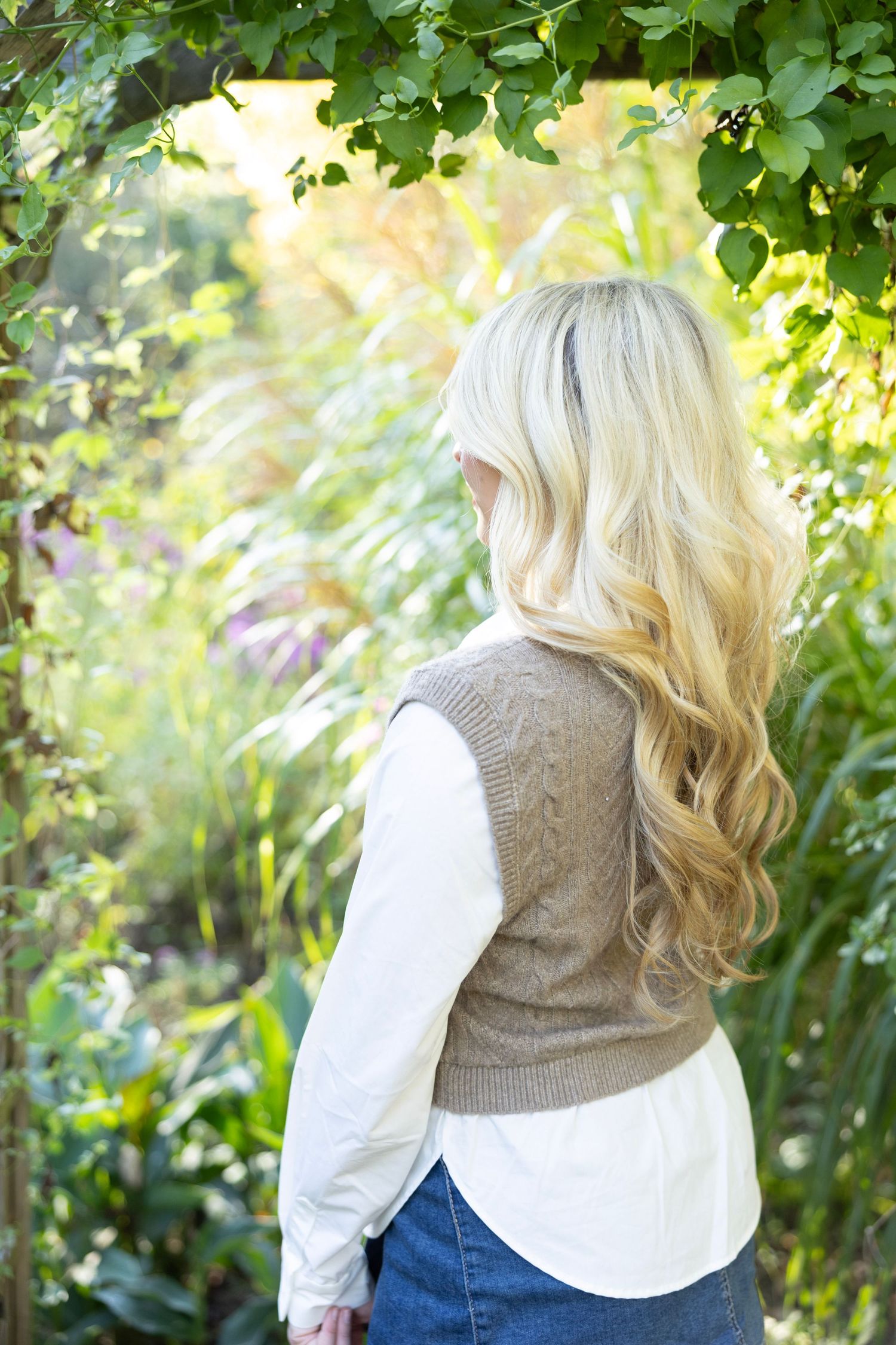 Back view portrait showing wavy blonde hair and knit vest against lush garden greenery.
