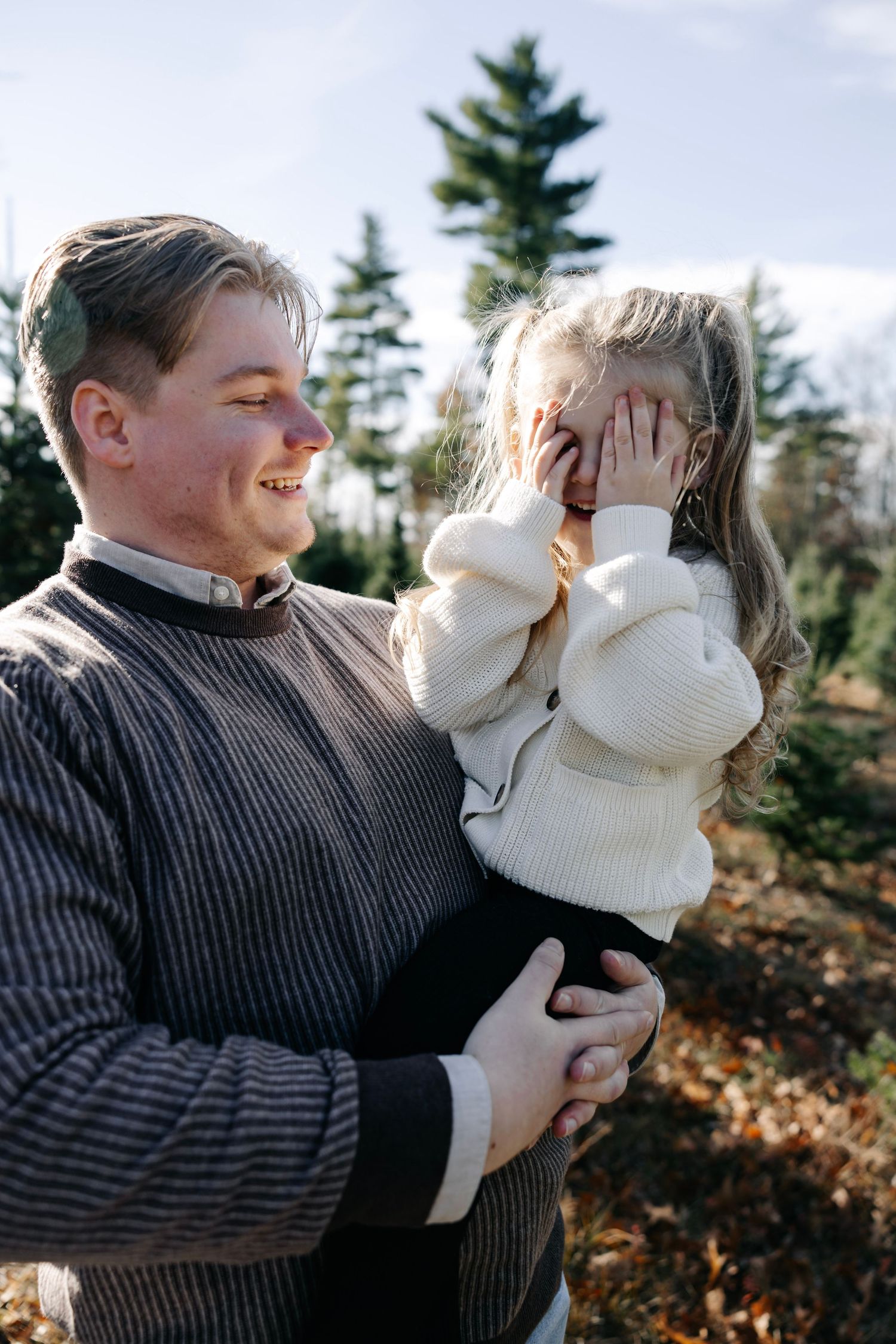 Playful moment between family members at Christmas tree farm with evergreens in background.