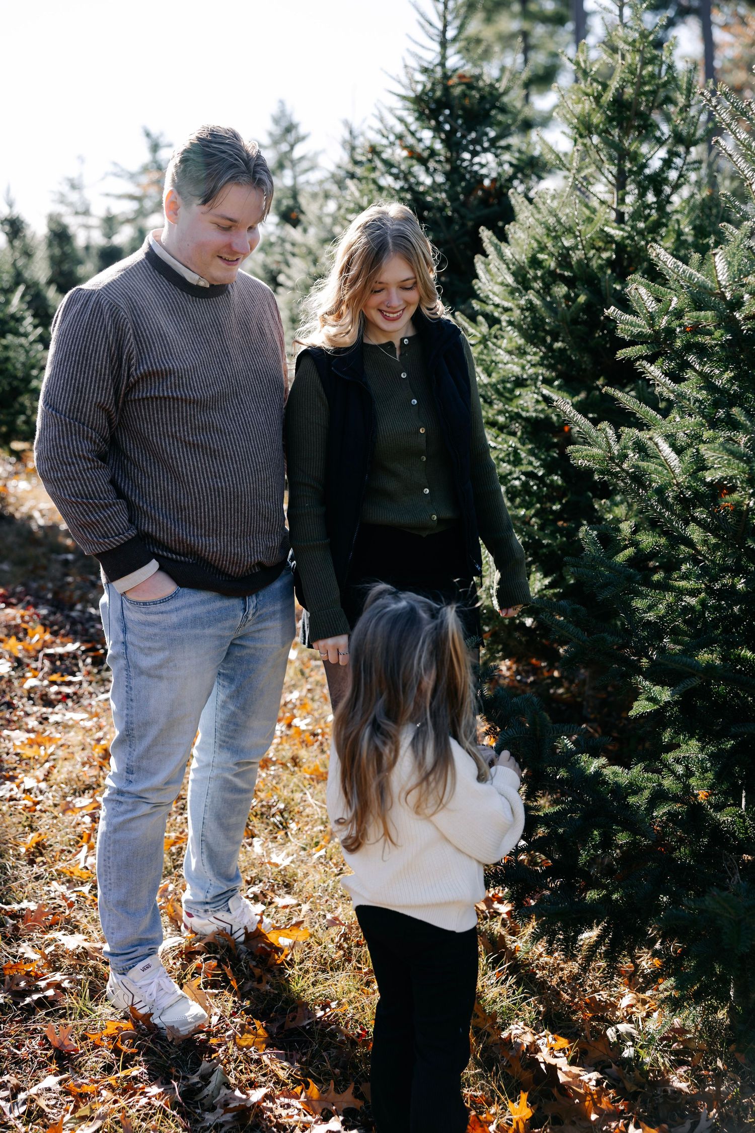 Casual family portrait among evergreen trees with fallen autumn leaves on the ground.