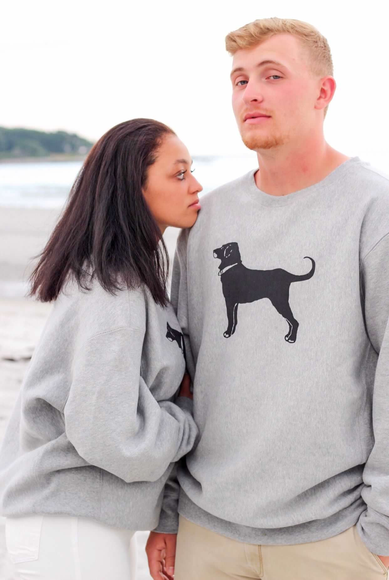A couple wearing matching gray sweatshirts with black dog designs pose together on a beach.