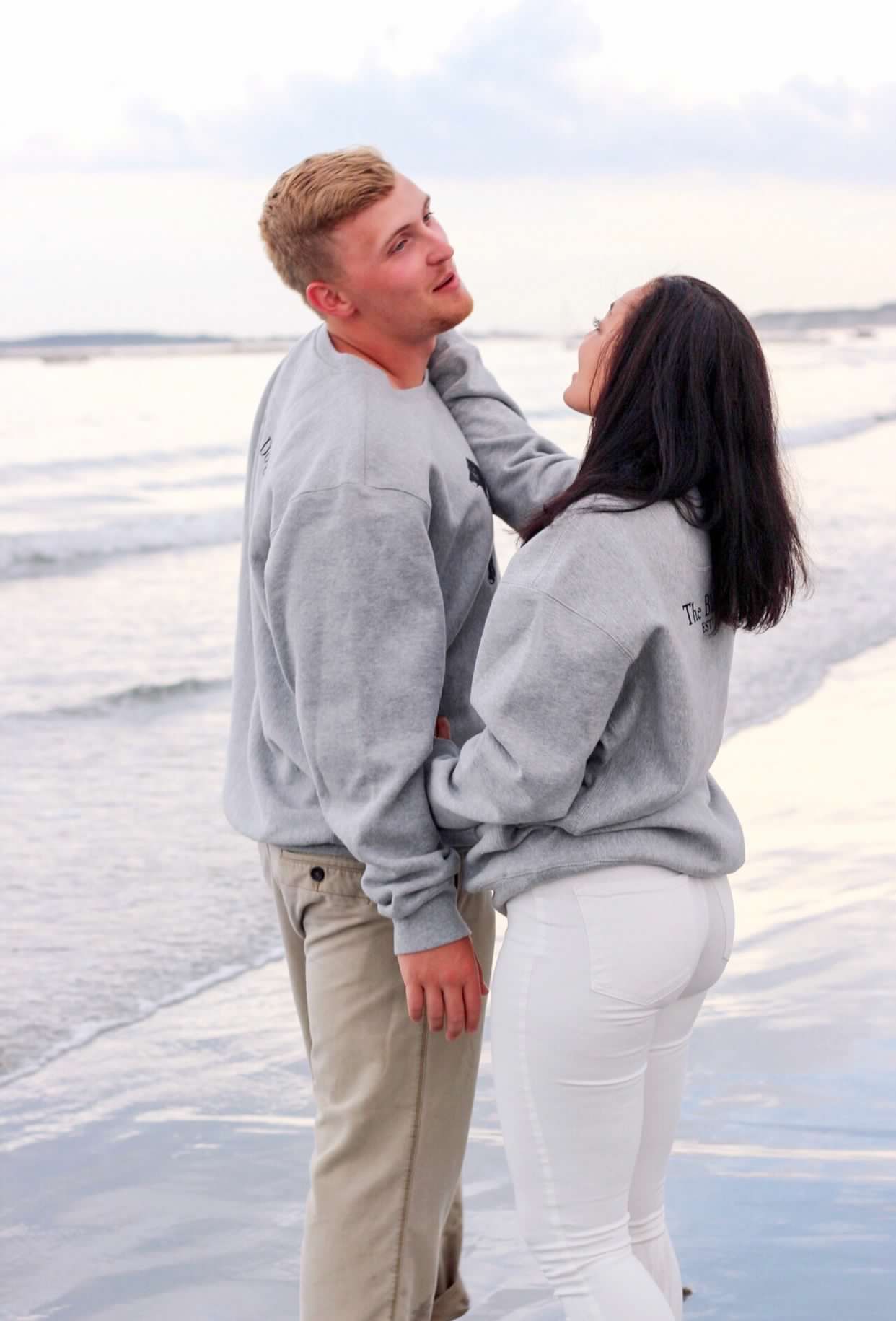 Couple in matching grey sweatshirts embrace on beach at sunset.