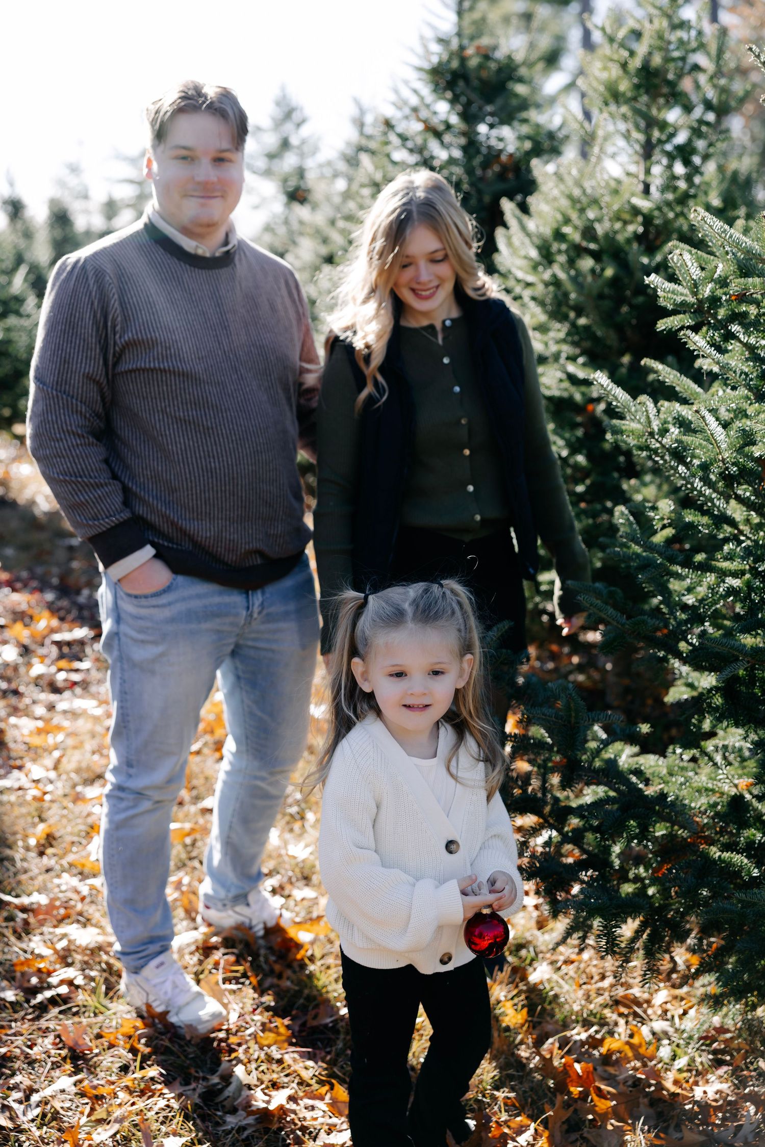 Family spending time together at a tree farm surrounded by evergreens and fallen leaves.