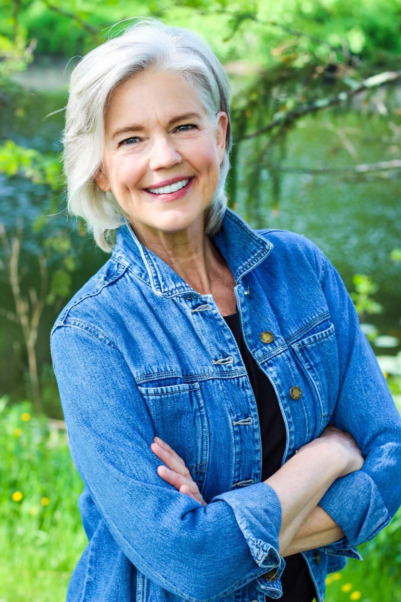 Person wearing denim jacket smiles warmly while posing in lush garden setting.