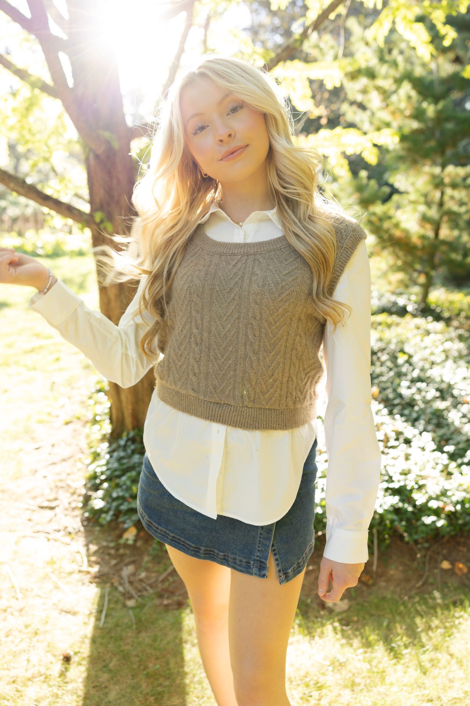 Person wearing brown cable knit sweater and white shirt poses in sunny garden setting.