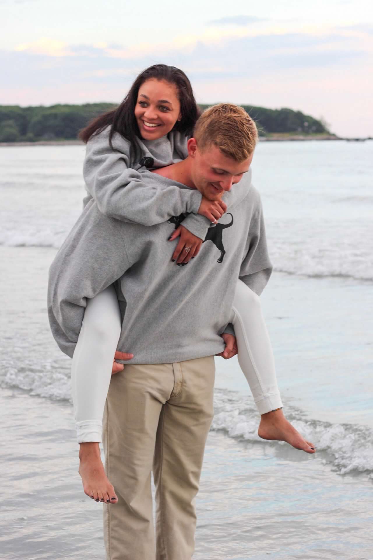 Playful couple having fun on the beach with piggyback ride in casual gray and white clothing.