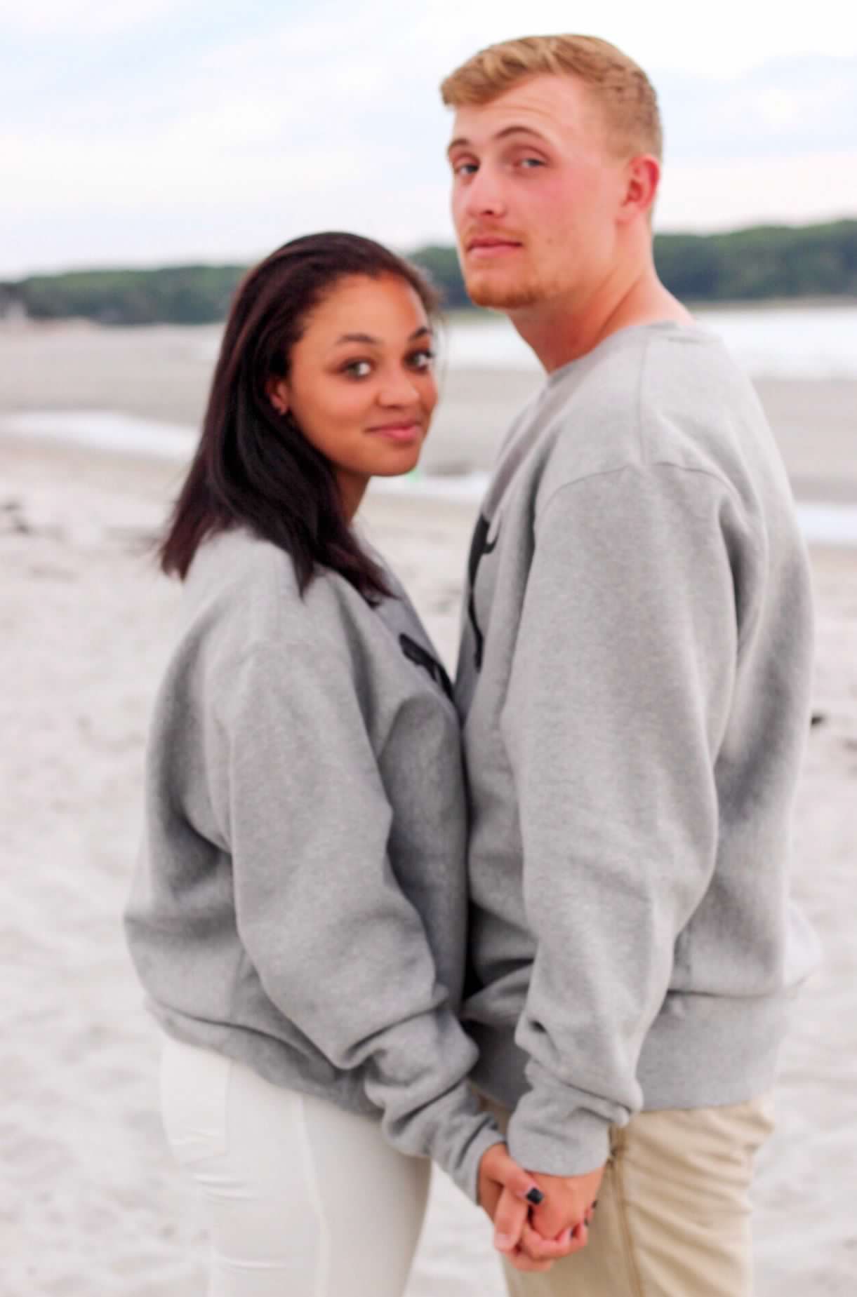 A couple in coordinated gray sweatshirts share an intimate moment on a sandy beach.