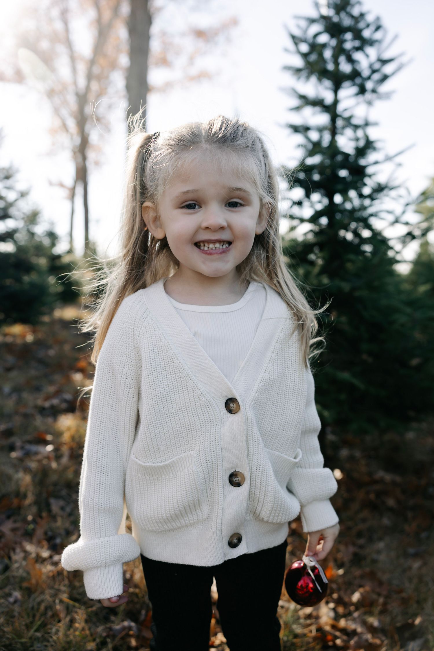 Child wearing white cardigan sweater smiling outdoors among evergreen trees.