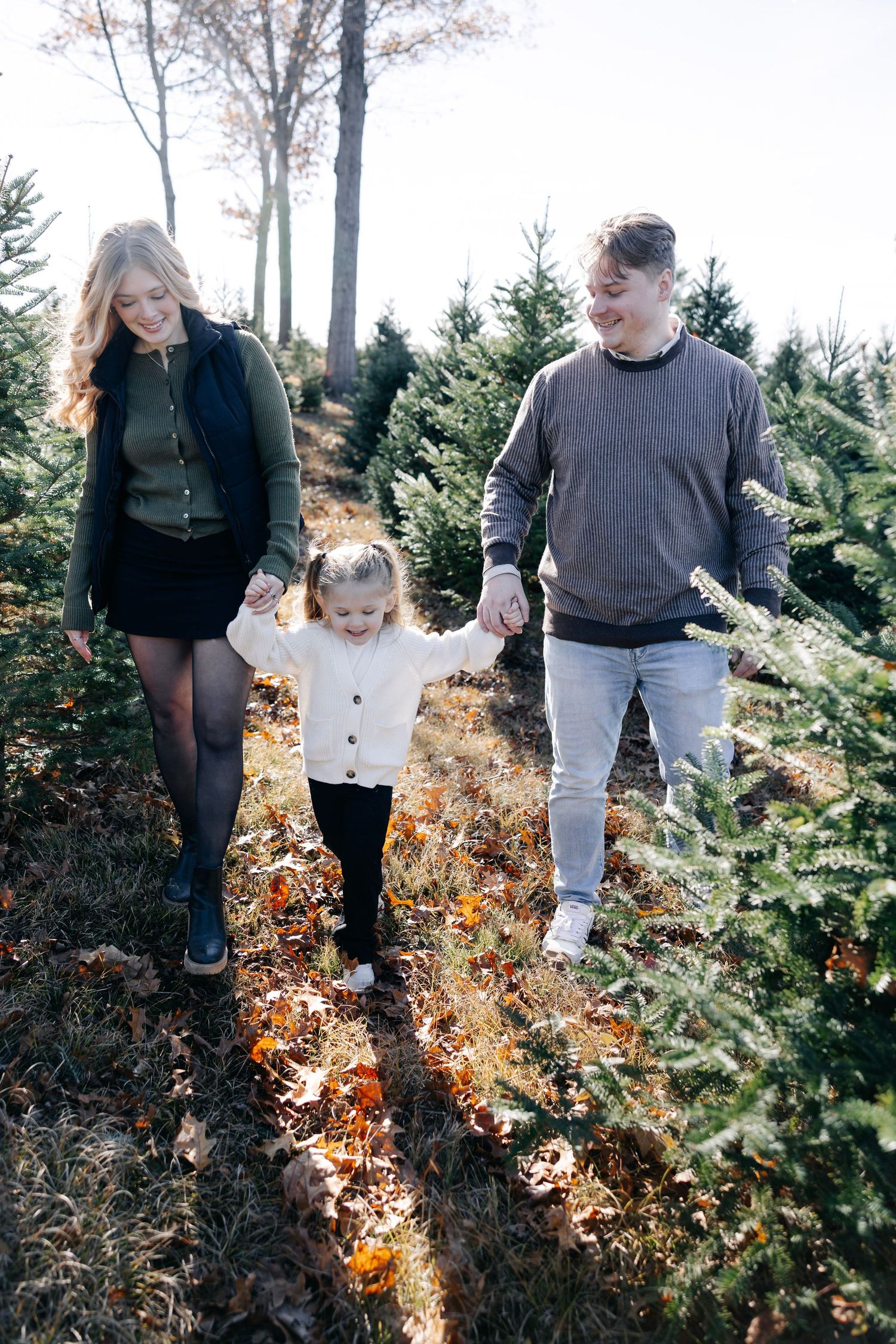 Family walking hand in hand through rows of Christmas trees on sunny afternoon.