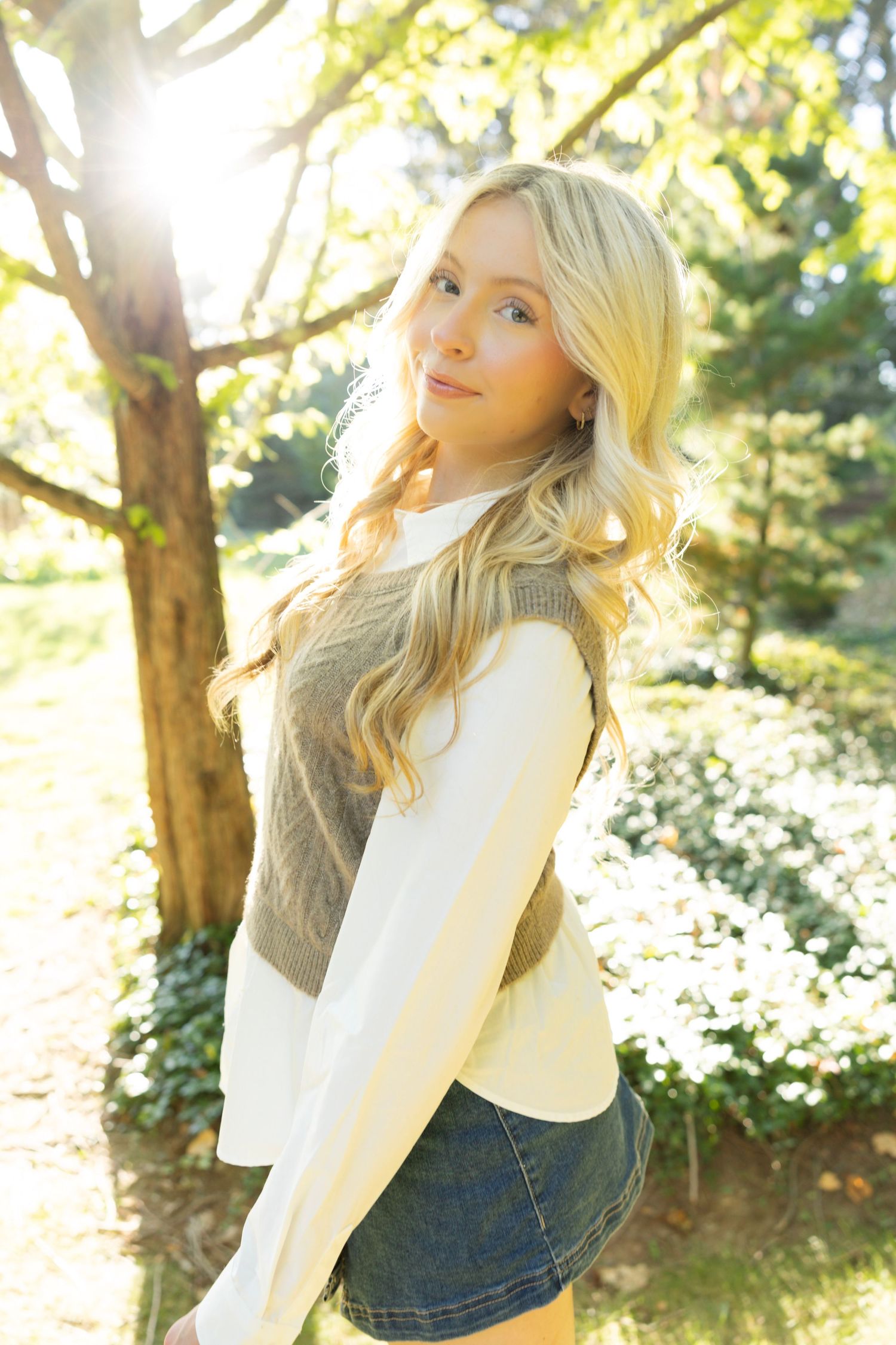 A fall fashion portrait in dappled sunlight featuring a knit sweater vest layered over white shirt and denim skirt.