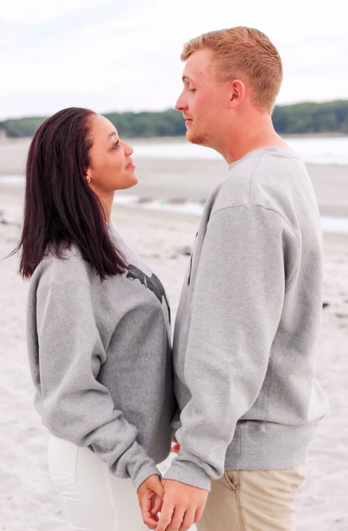 A romantic couple in gray sweatshirts hold hands while standing on a beach.