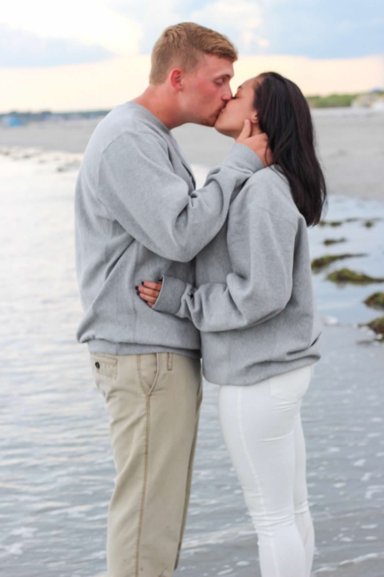 Romantic couple embracing on a beach at sunset wearing matching gray and white outfits.
