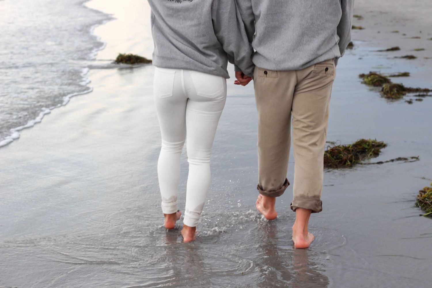 Two people walking barefoot together in shallow beach water, wearing white and khaki pants.