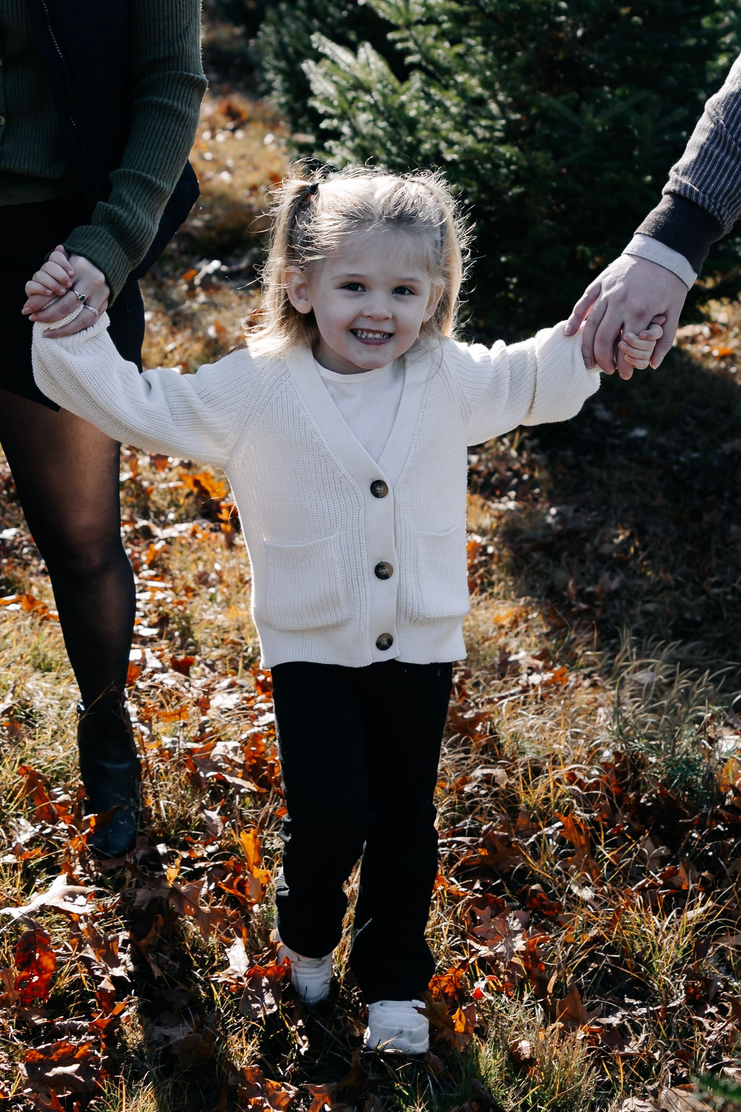 Young child in white sweater holding hands with adults while walking through autumn foliage.