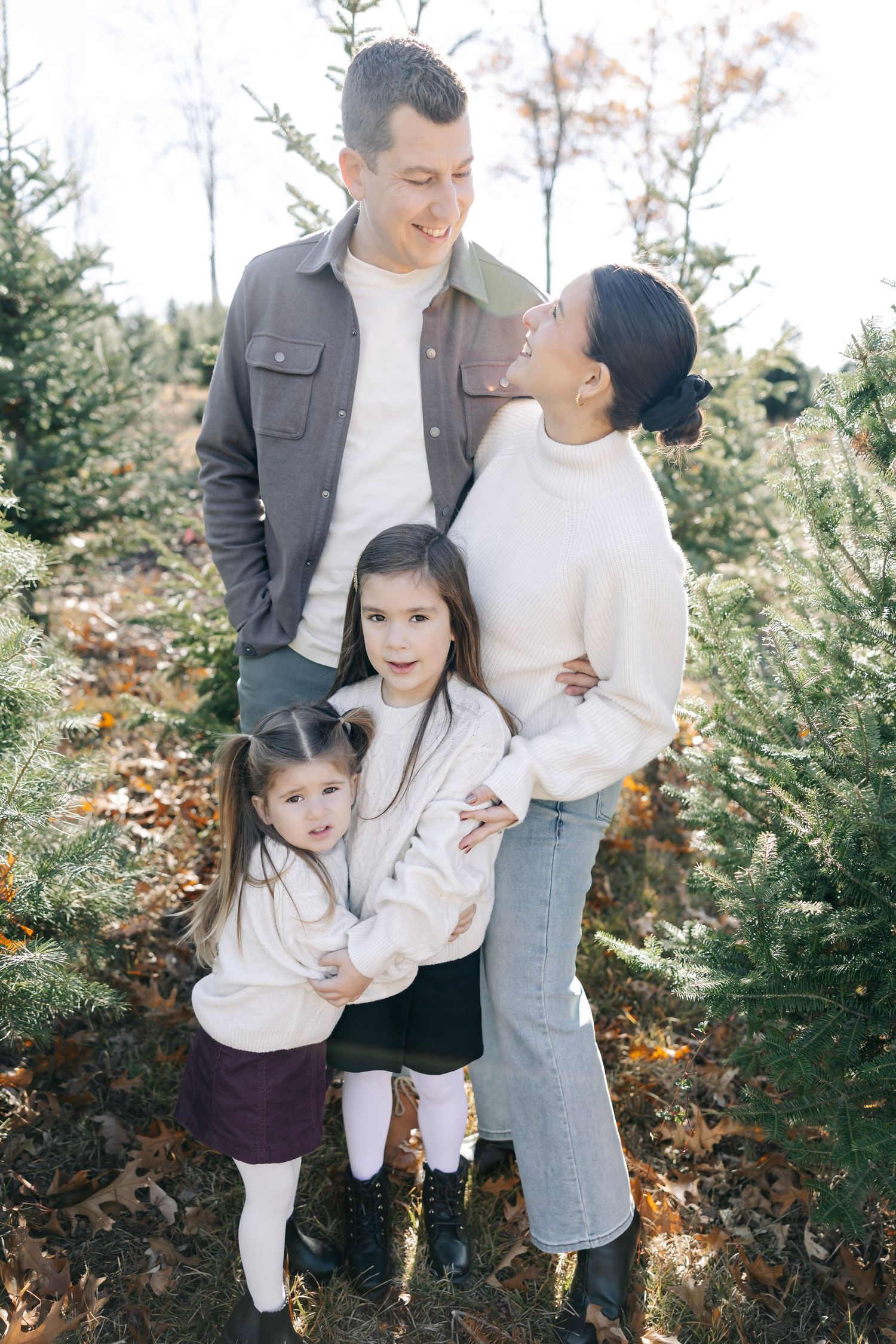 A family in coordinating white and grey outfits poses together among evergreen trees on an autumn day.
