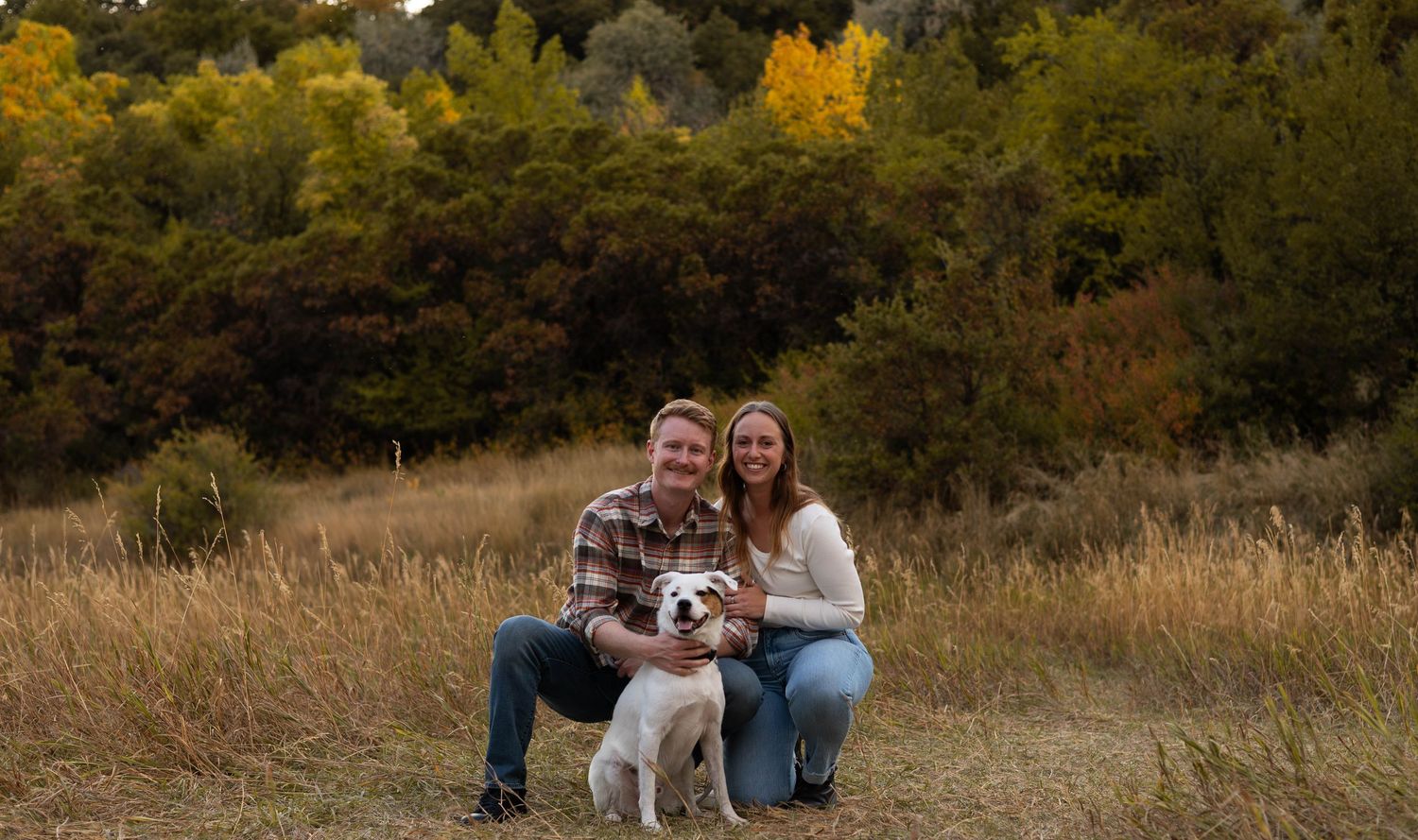 a couple kneeling down with their dog and smiling at the camera. there background has trees with fall colors.