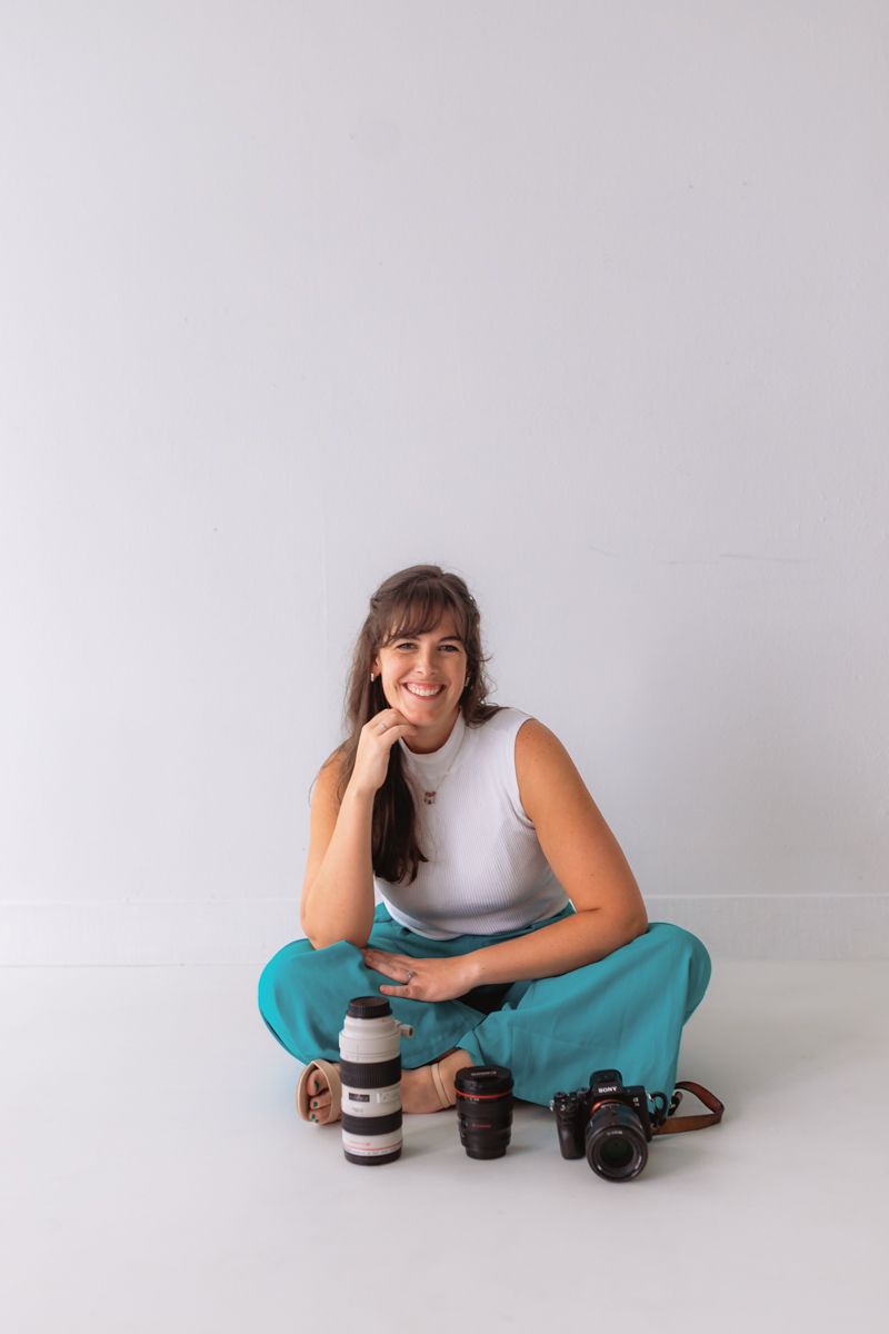 A person in a white tank top and teal pants sits on a gray floor with camera equipment and coffee cups.