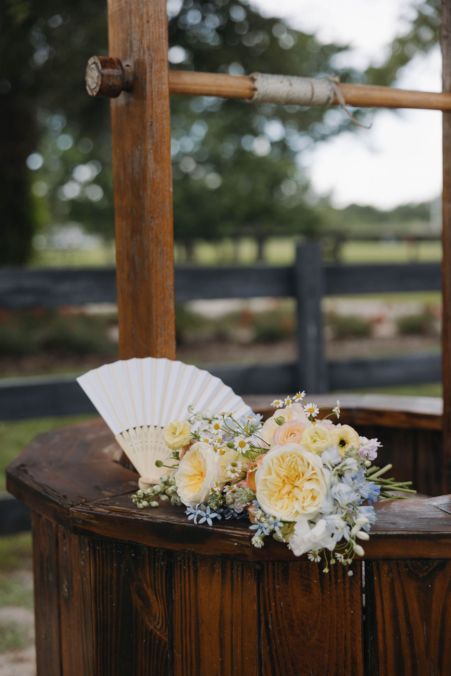 A bridal bouquet of soft yellow and white roses with blue accents rests beside a white fan on a rustic wooden stand.