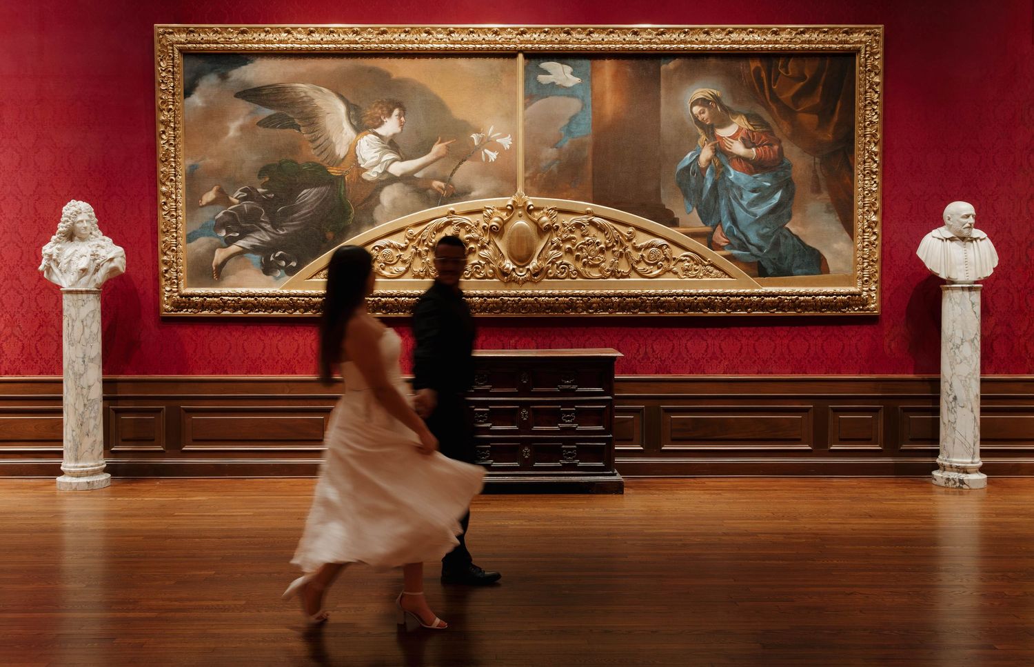 Two visitors walk past a large classical painting in a grand red-walled gallery with marble busts on pedestals.