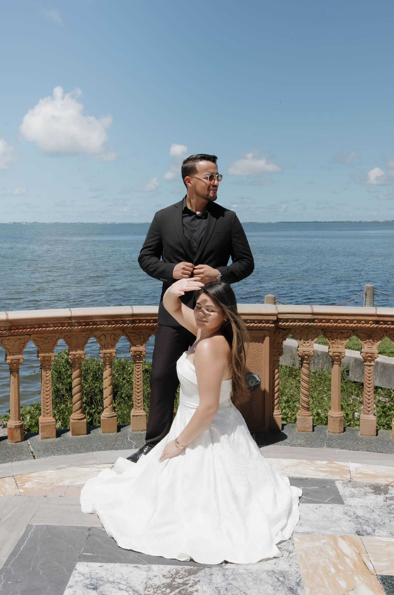 Bride in white gown sits before groom in black tuxedo on ornate terrace overlooking blue ocean under sunny sky.