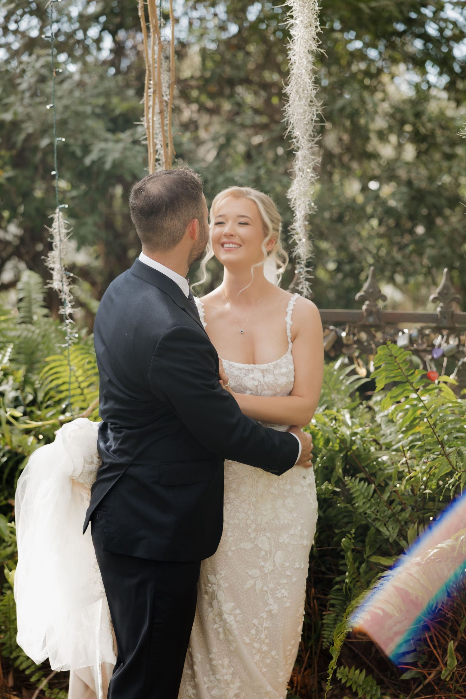 A couple shares a joyful moment together outdoors surrounded by lush ferns and hanging decorations at their wedding.