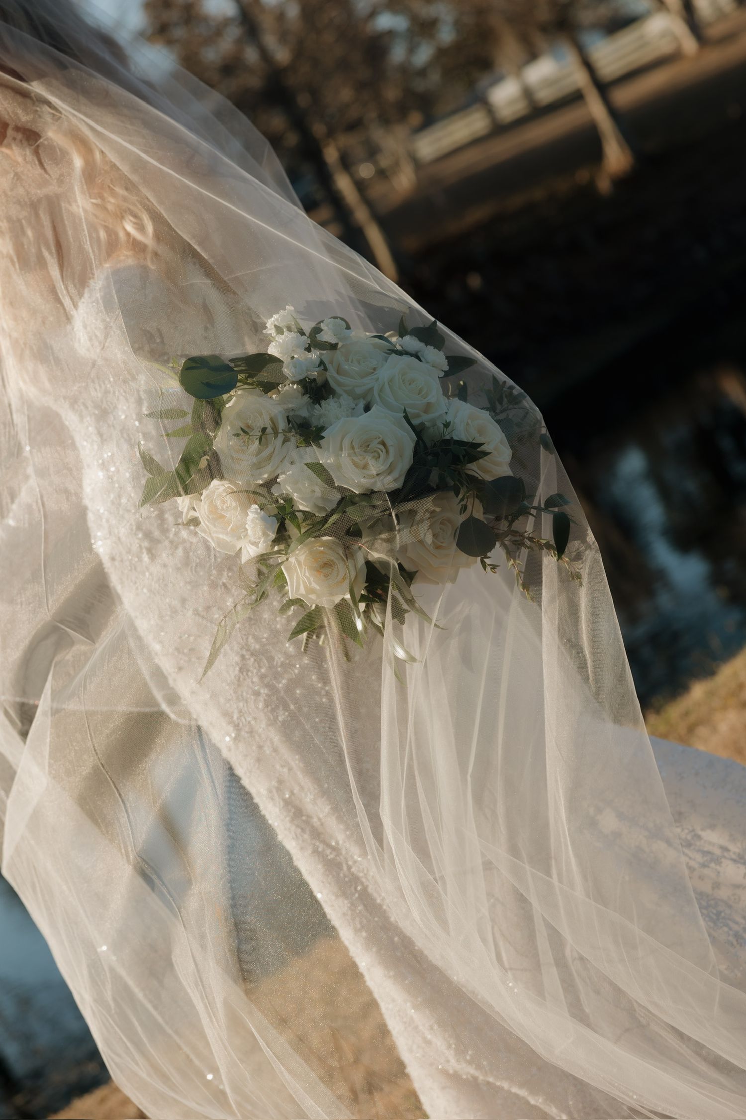 Bride holding white rose bouquet draped under flowing wedding veil outdoors near a bridge.