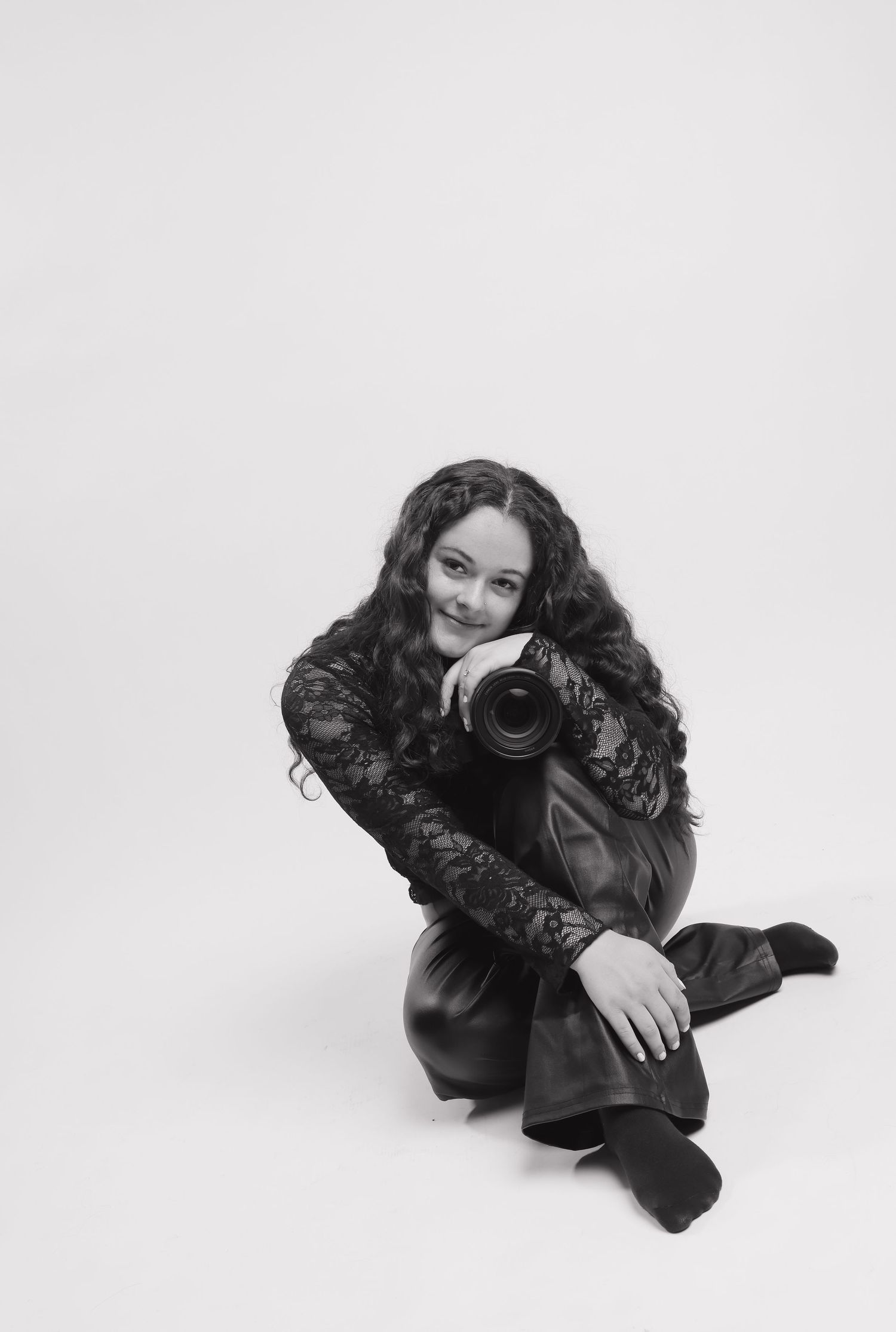 Black and white photo of a smiling woman with curly hair holding a camera, sitting on a white studio floor.