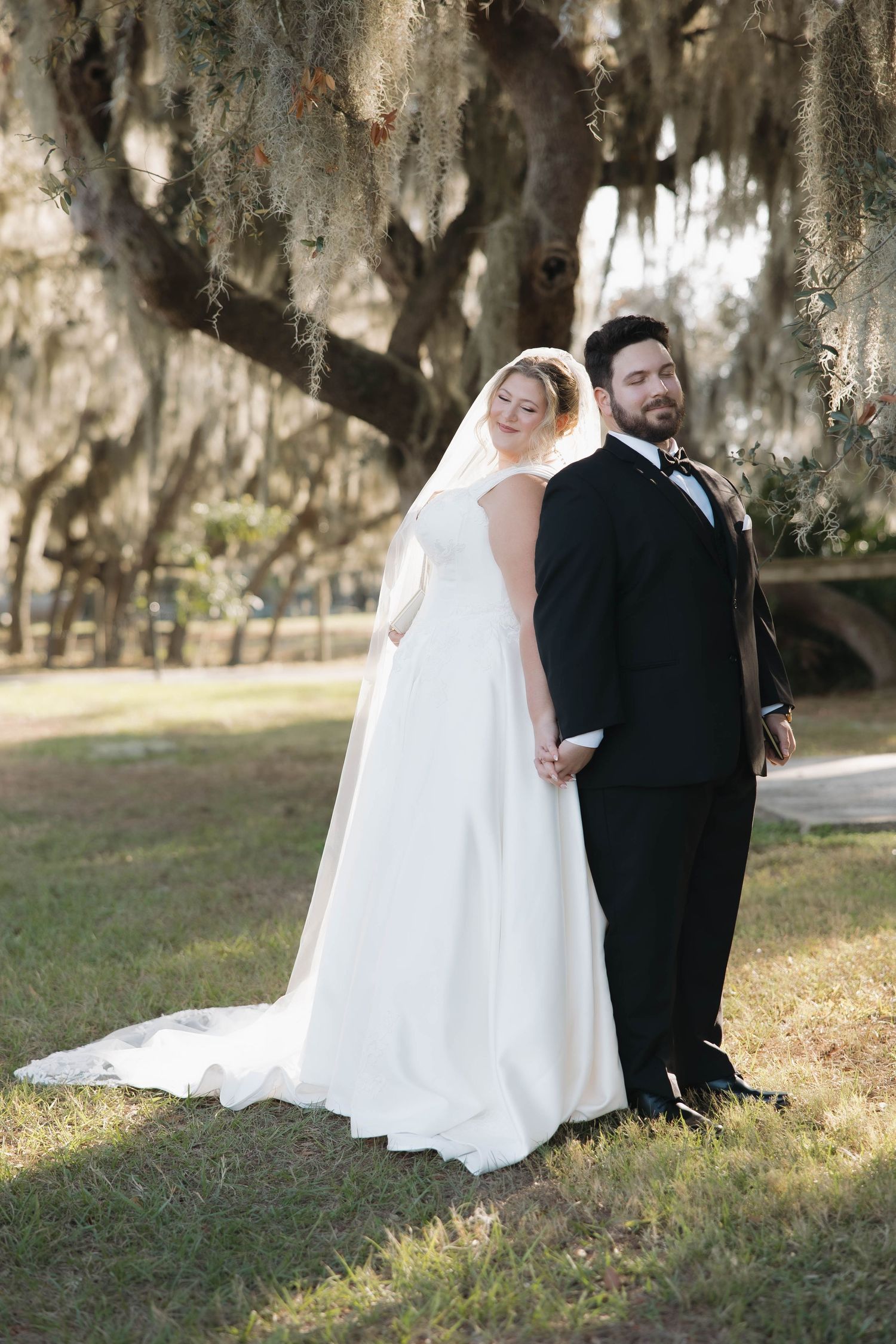 A bride and groom pose together under Spanish moss draped trees in a romantic outdoor wedding setting.