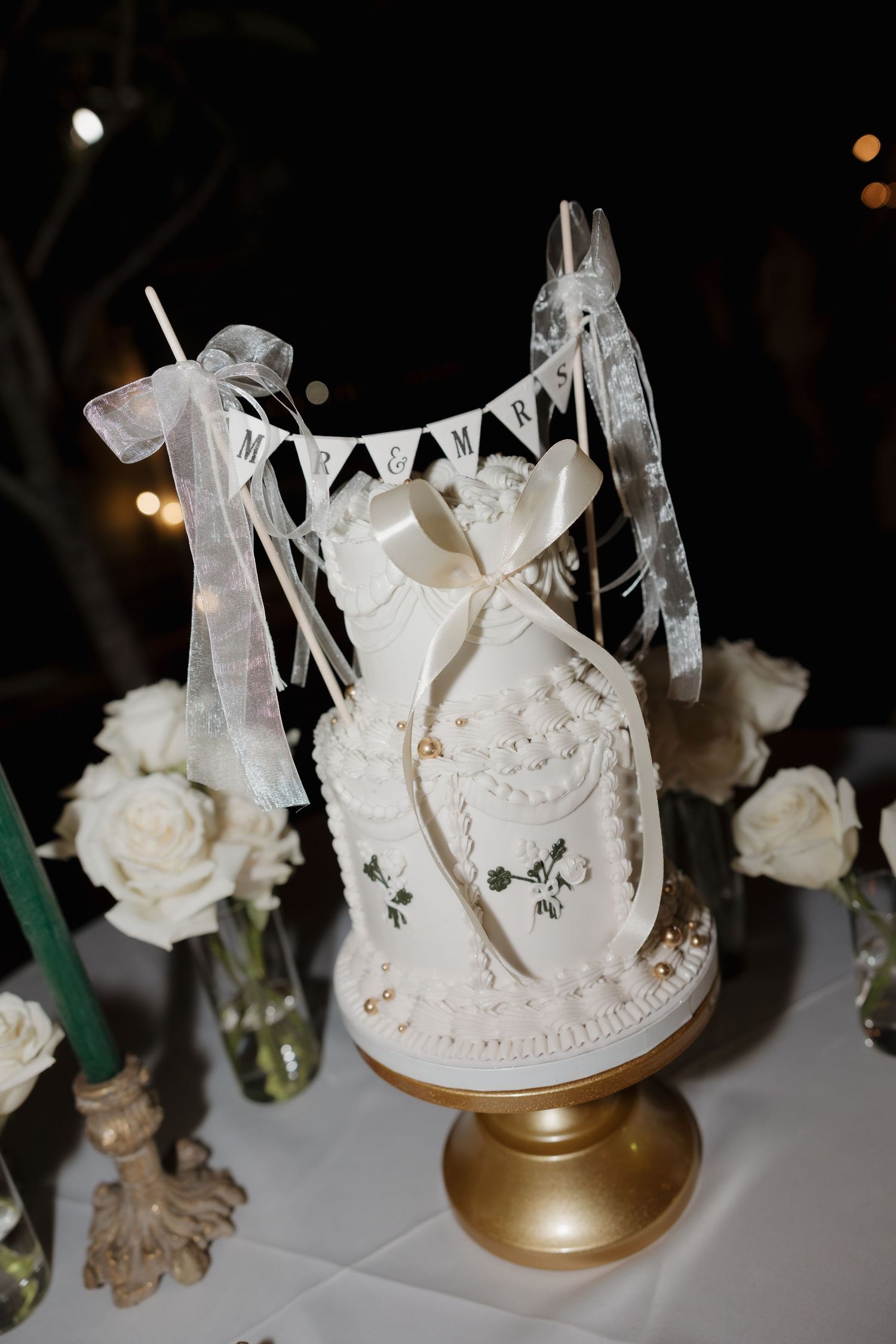 White wedding cake topper with delicate details sits atop a gold pedestal surrounded by white roses.