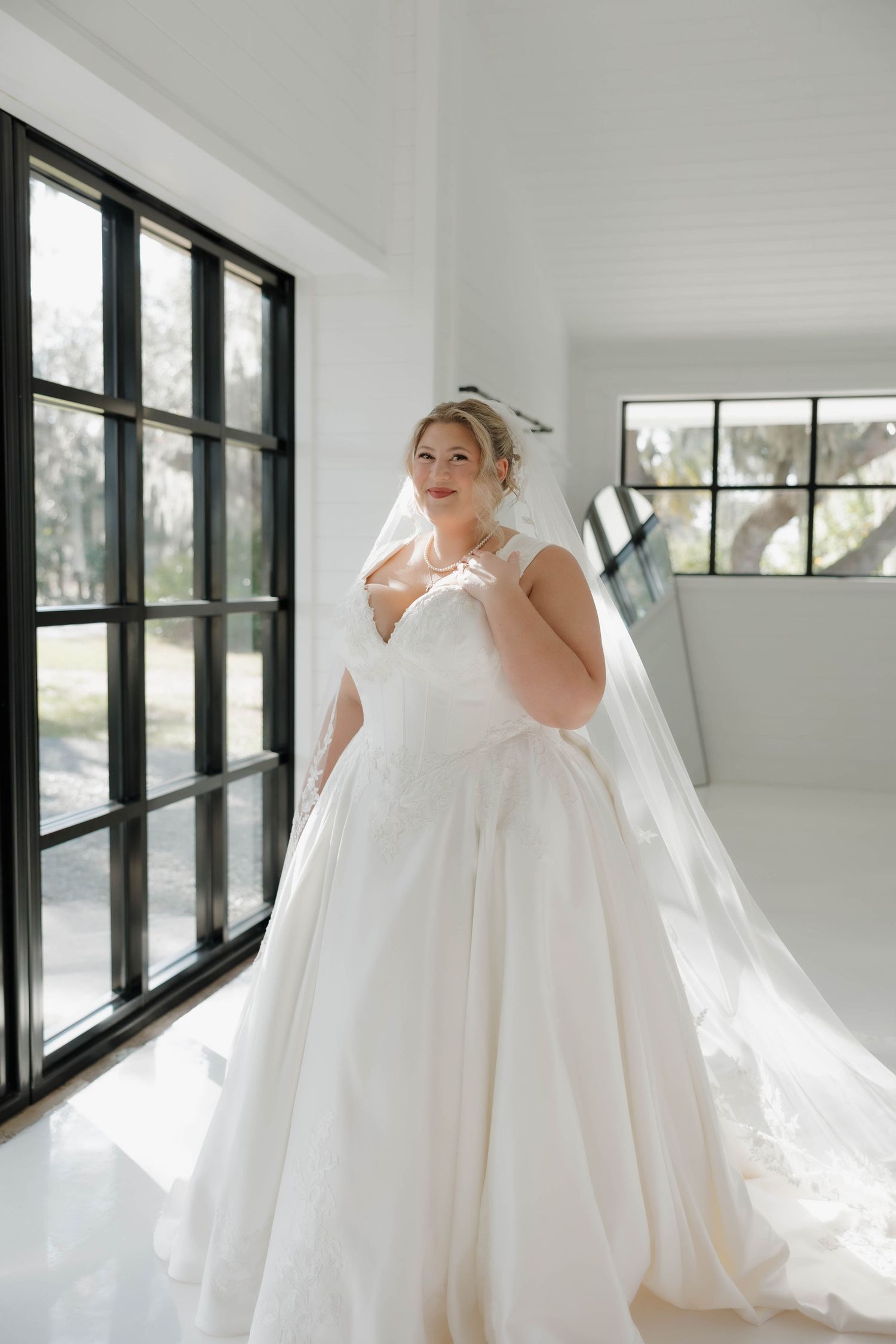 A bride in a flowing white wedding gown stands in a bright sunlit room with black-framed windows.