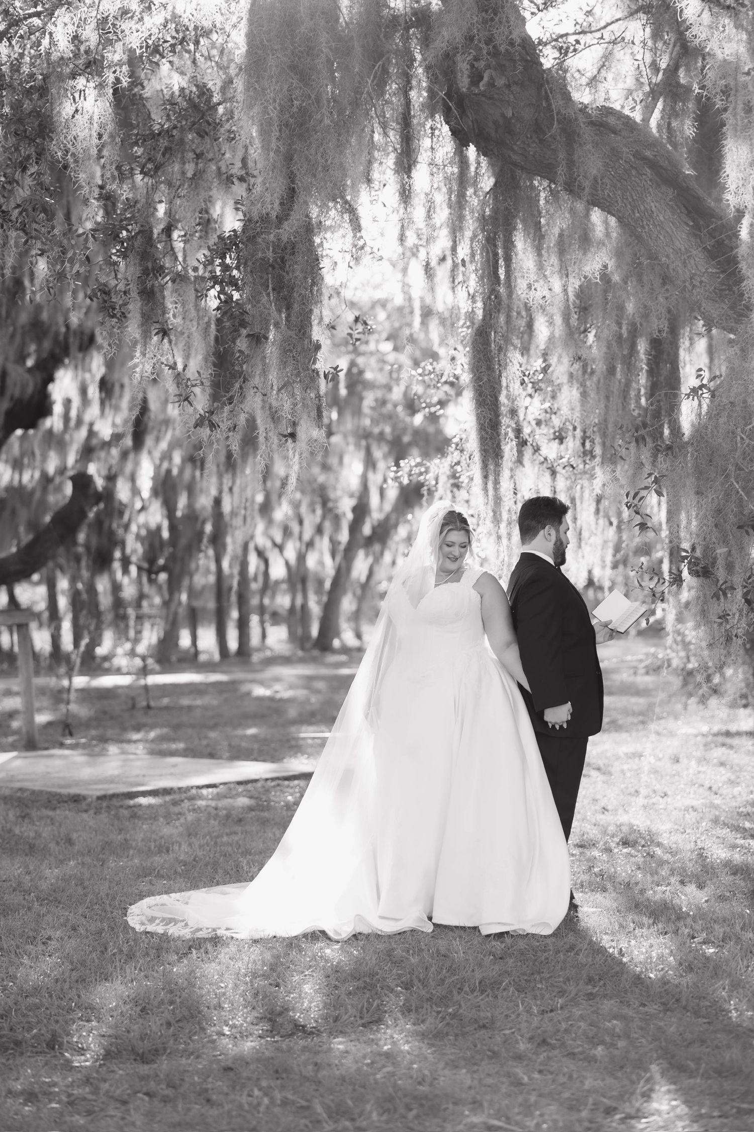 Black and white photo of a bride and groom walking hand in hand under Spanish moss-draped oak trees.