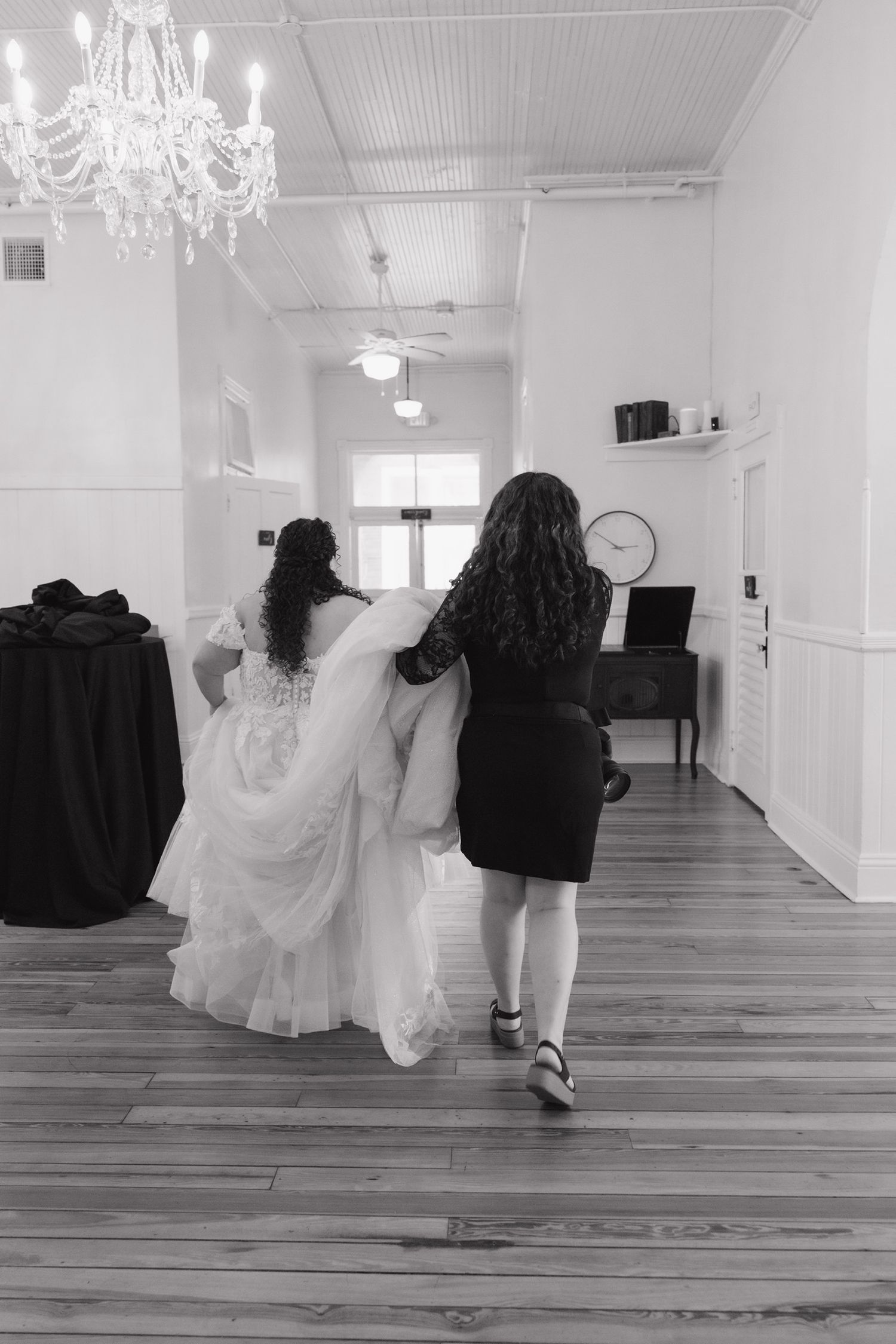 Black and white photo of bride in wedding gown being helped by assistant walking down hall with chandelier.