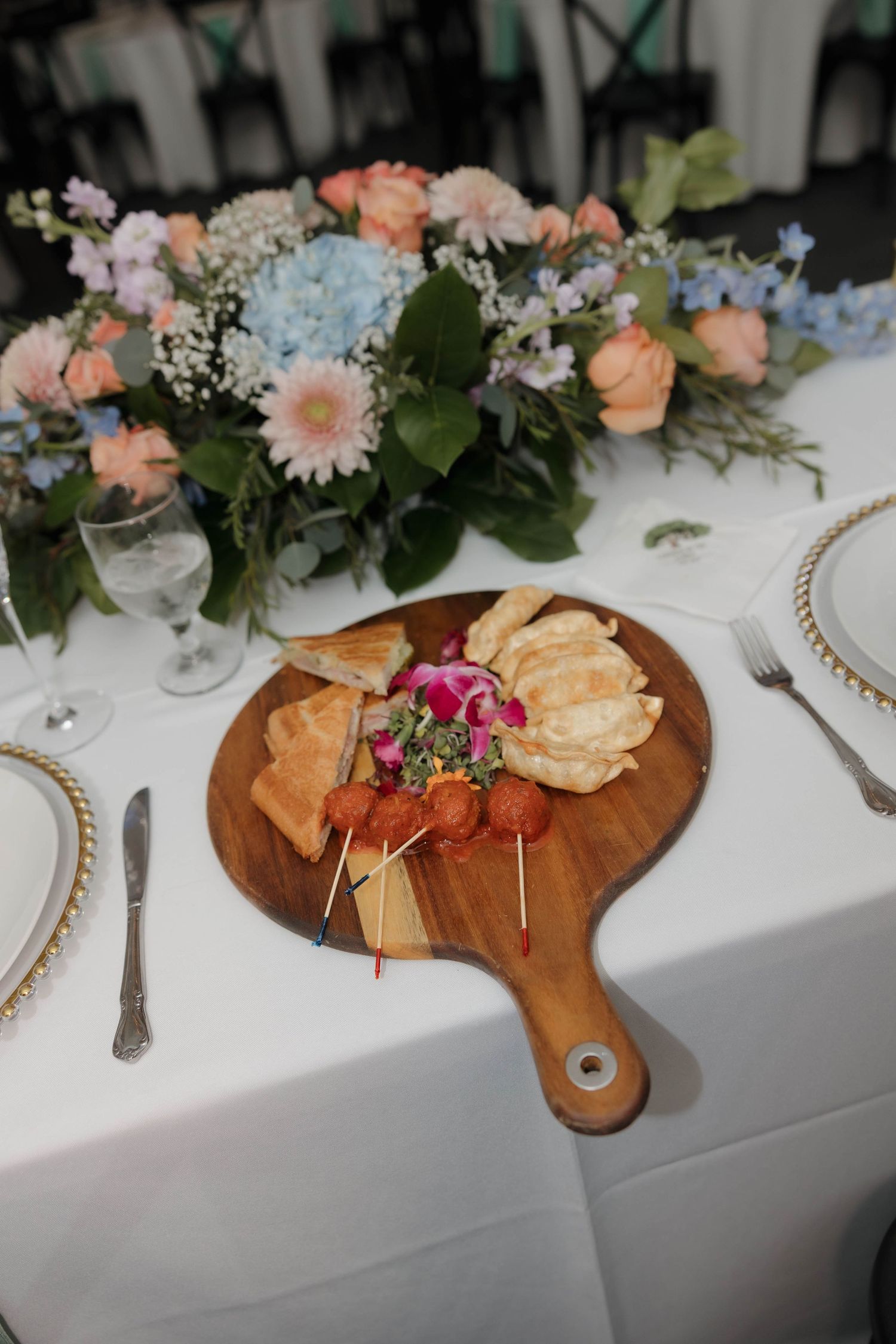 Wooden charcuterie boards with appetizers and snacks arranged on a white tablecloth with floral centerpieces.