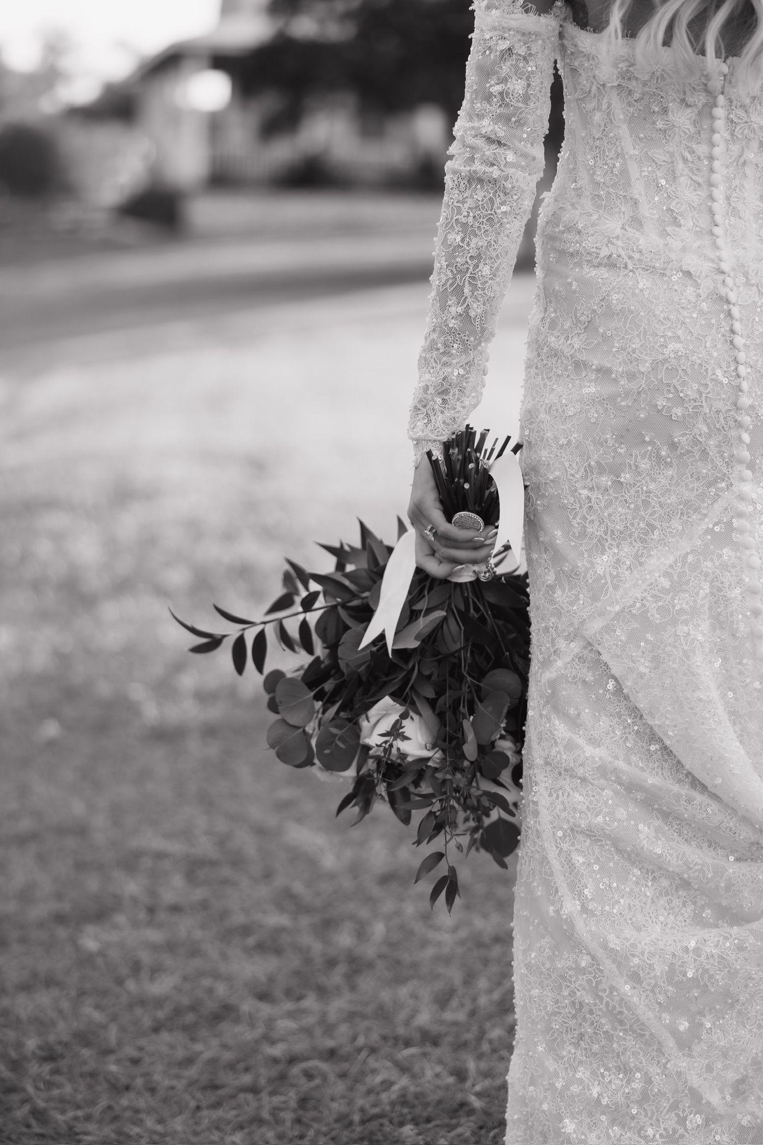 Black and white photo of a bride in a beaded lace gown holding a lush greenery bouquet outdoors.
