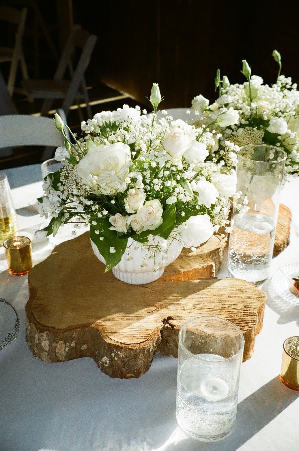 White roses and baby's breath centerpiece in white vase on rustic wood slice, elegant wedding table decor.