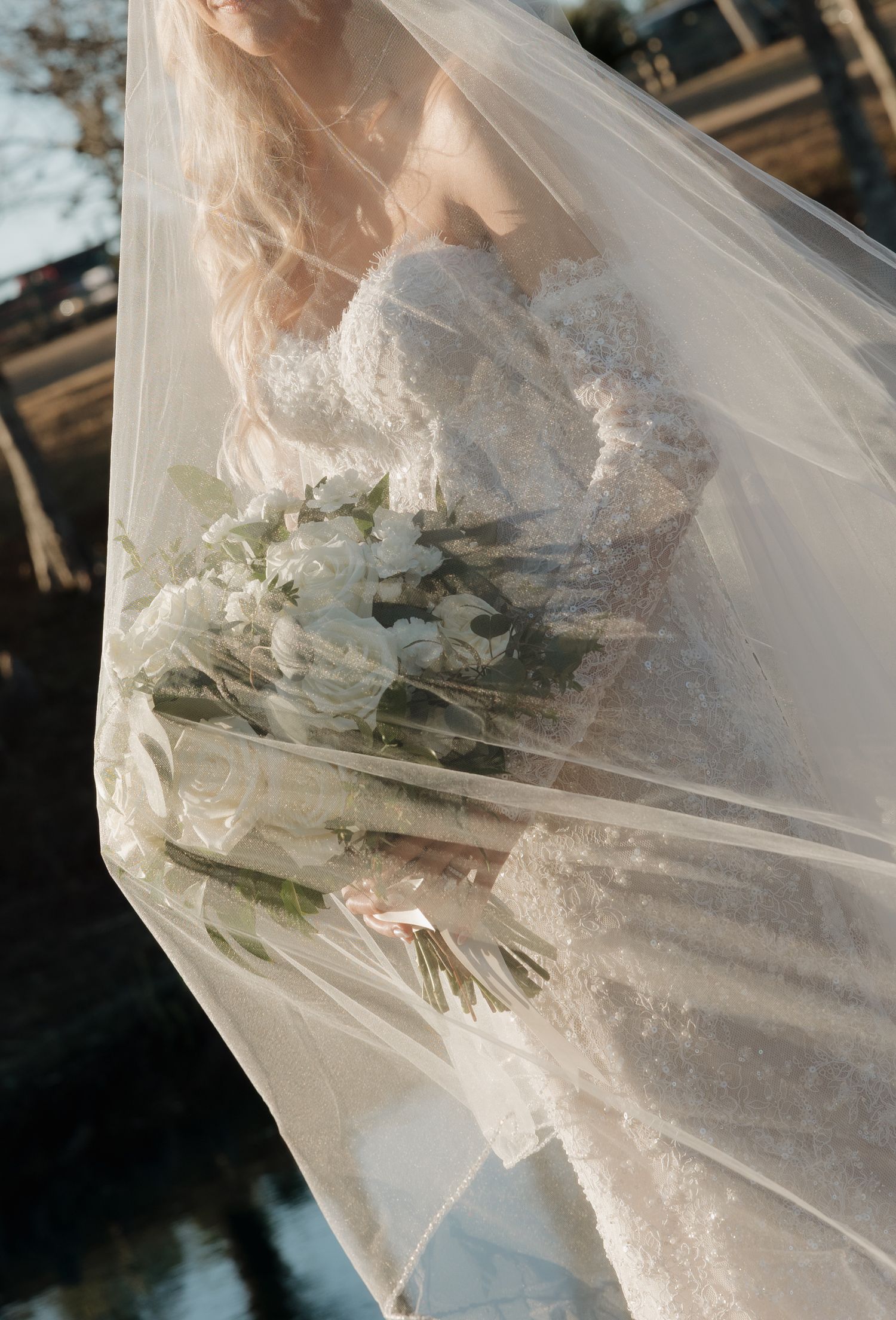 A bride in a delicate lace wedding dress holds a white bouquet under a flowing sheer veil in soft sunlight.
