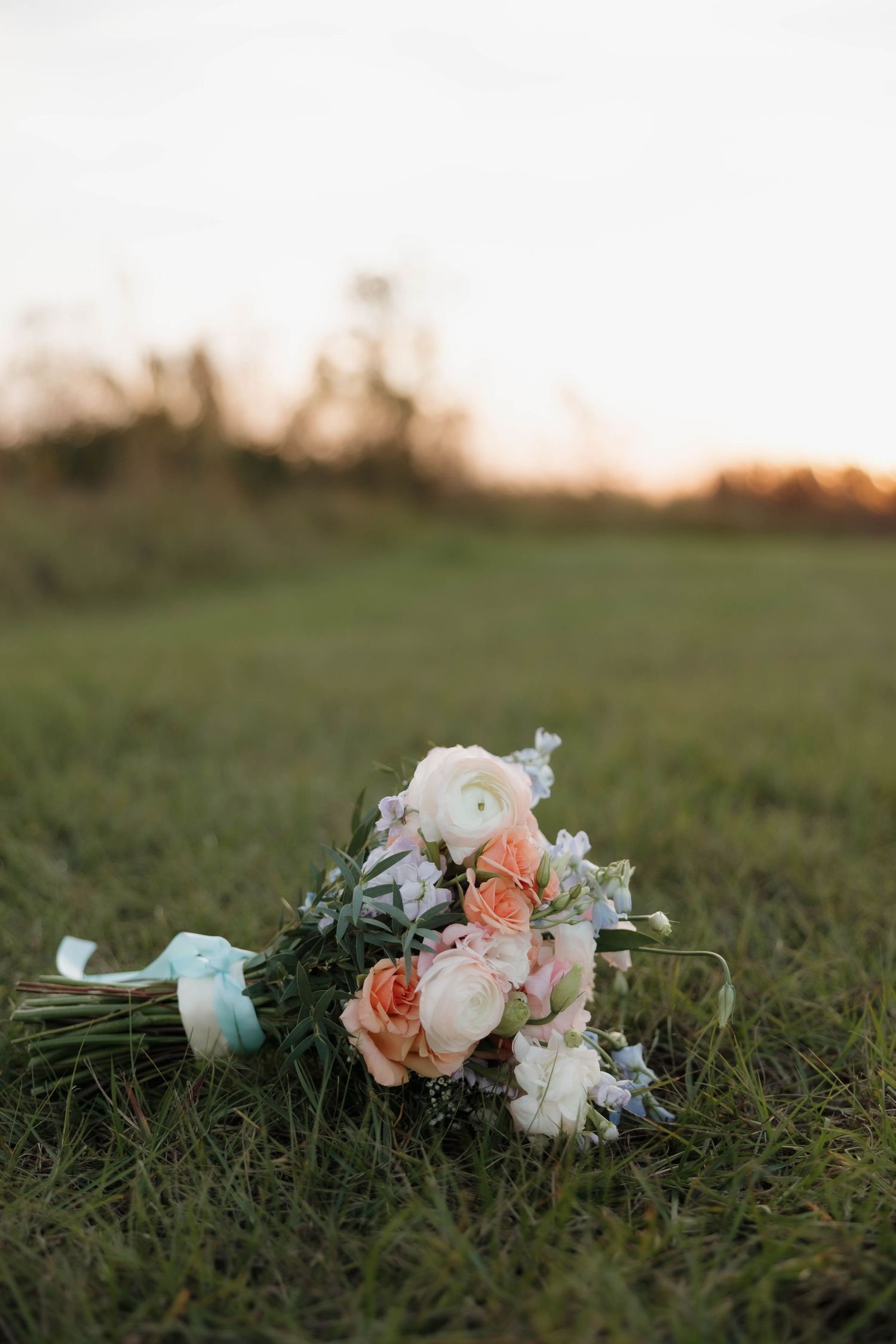 A delicate wedding bouquet with peach and white flowers tied with a blue ribbon rests on green grass at sunset.