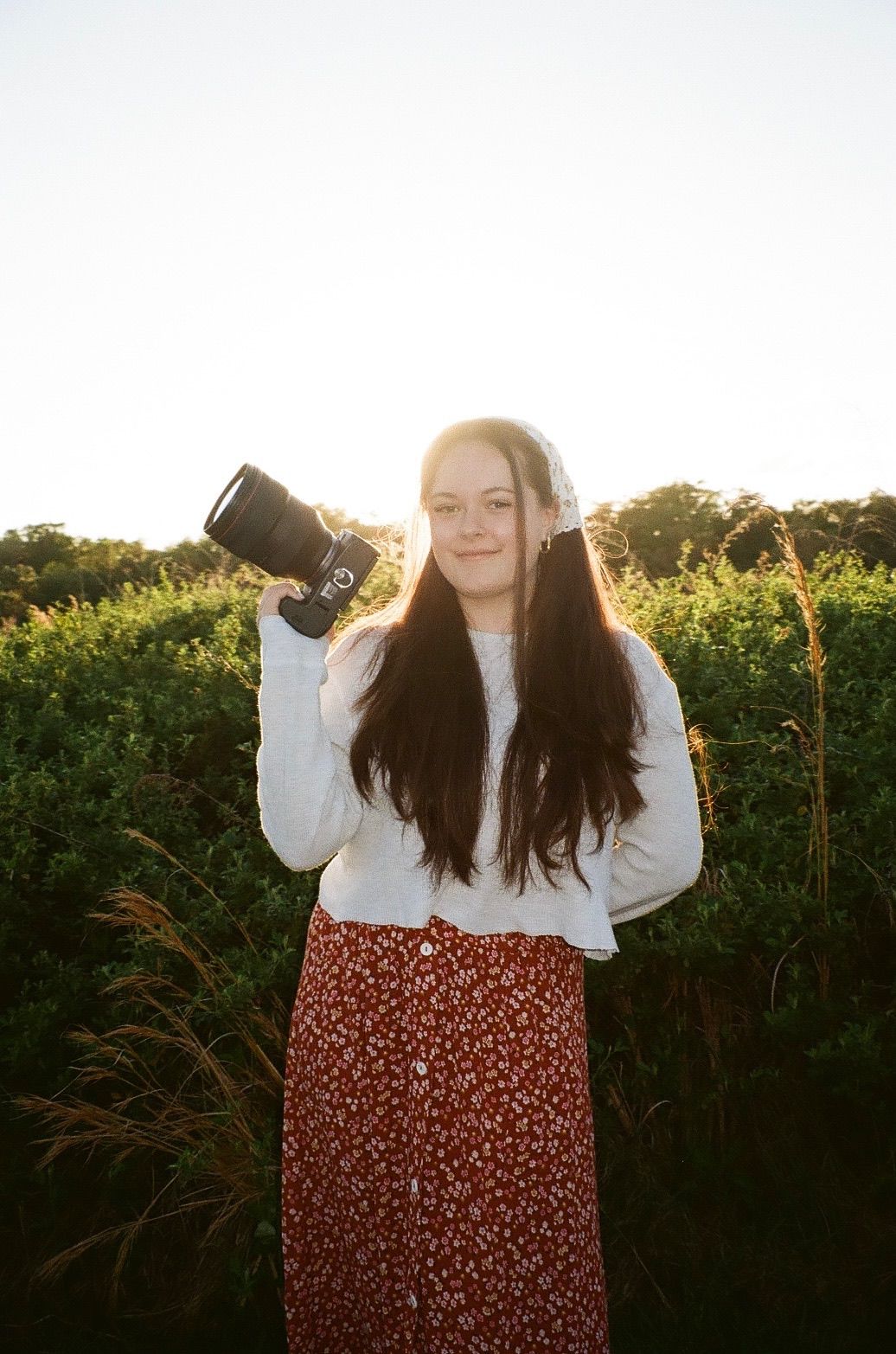 Young woman with long dark hair holding a camera, wearing a white top and floral skirt, standing in a sunlit green field.