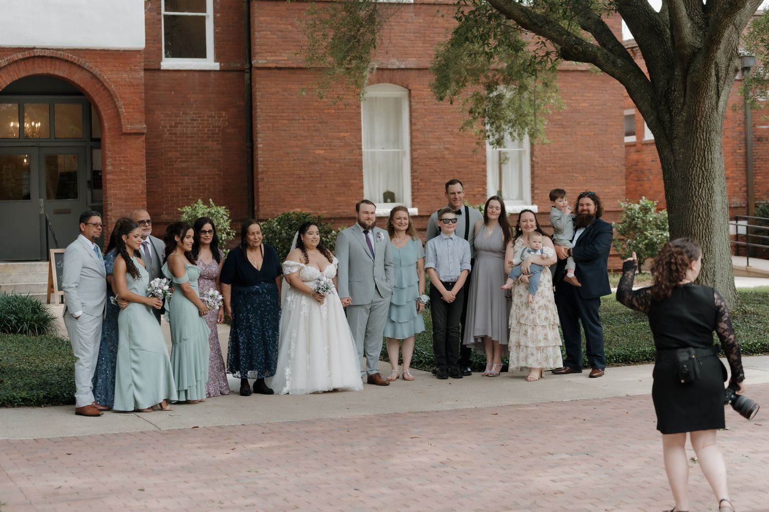 Wedding party poses for group photo outside brick building, bride in white gown surrounded by guests in sage green and gray.