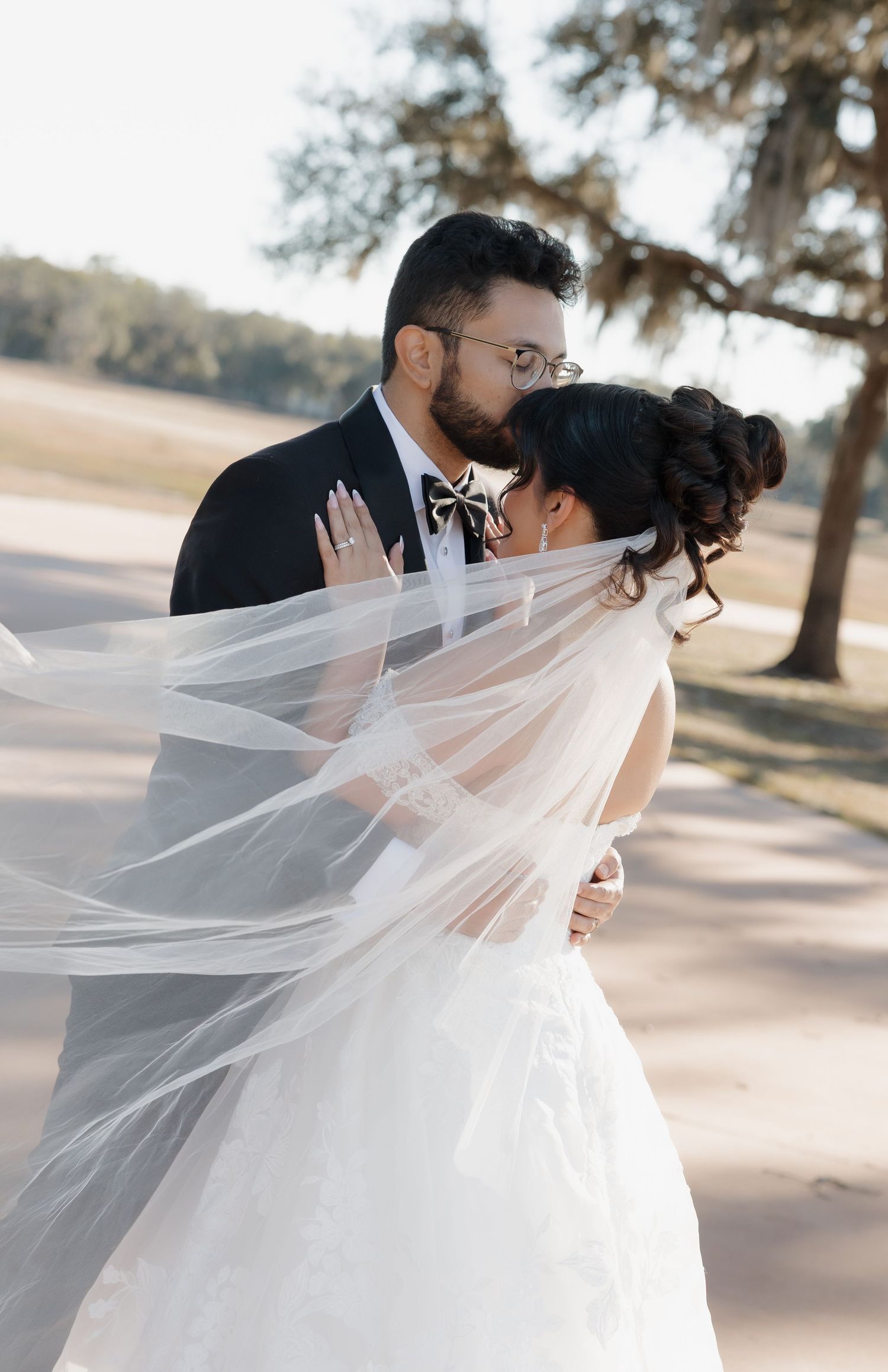 A romantic wedding couple shares a tender kiss as the bride's flowing white veil catches the breeze in golden sunlight.