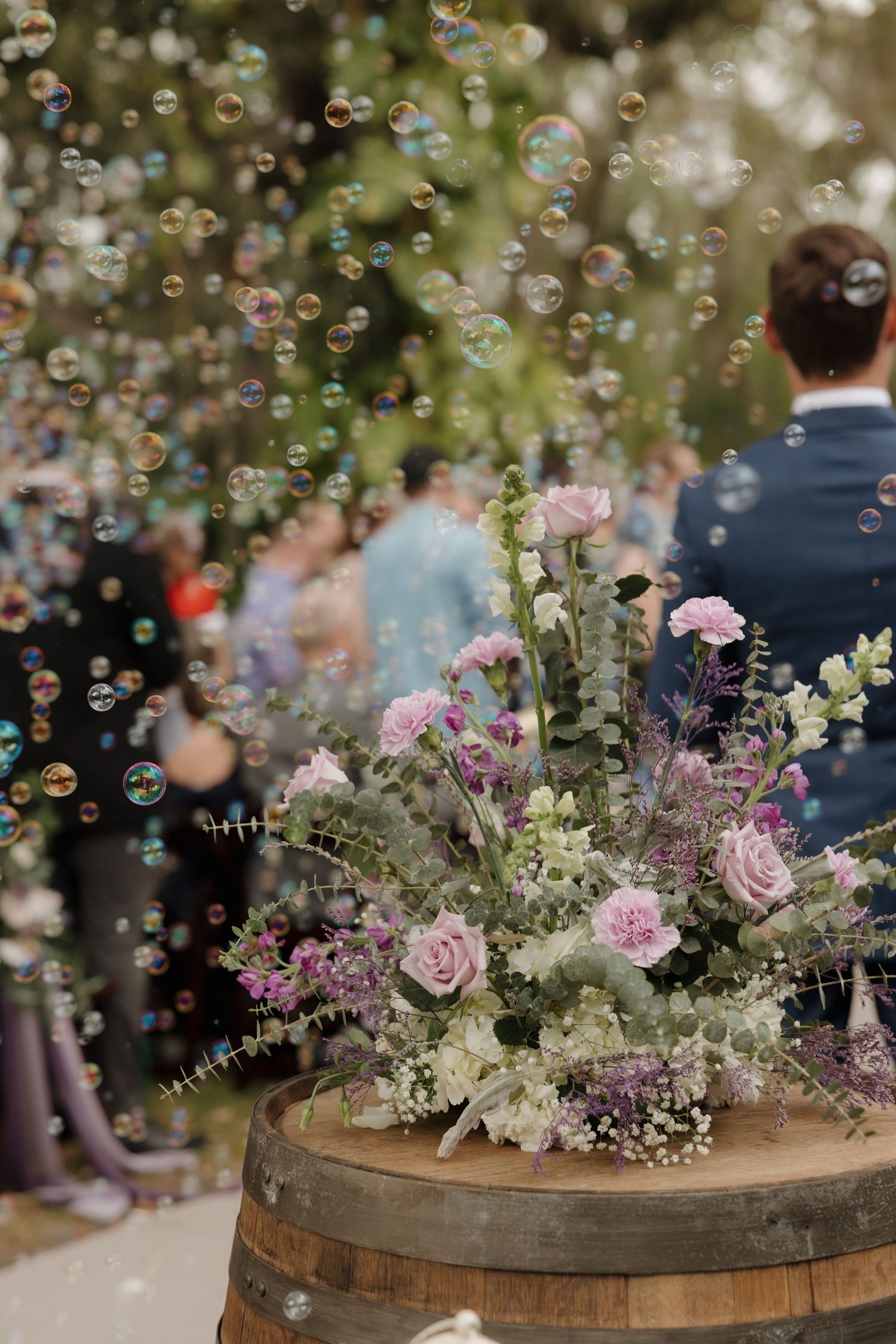 Floral arrangement of pink roses and greenery on a wooden barrel with bubbles floating at an outdoor wedding celebration.