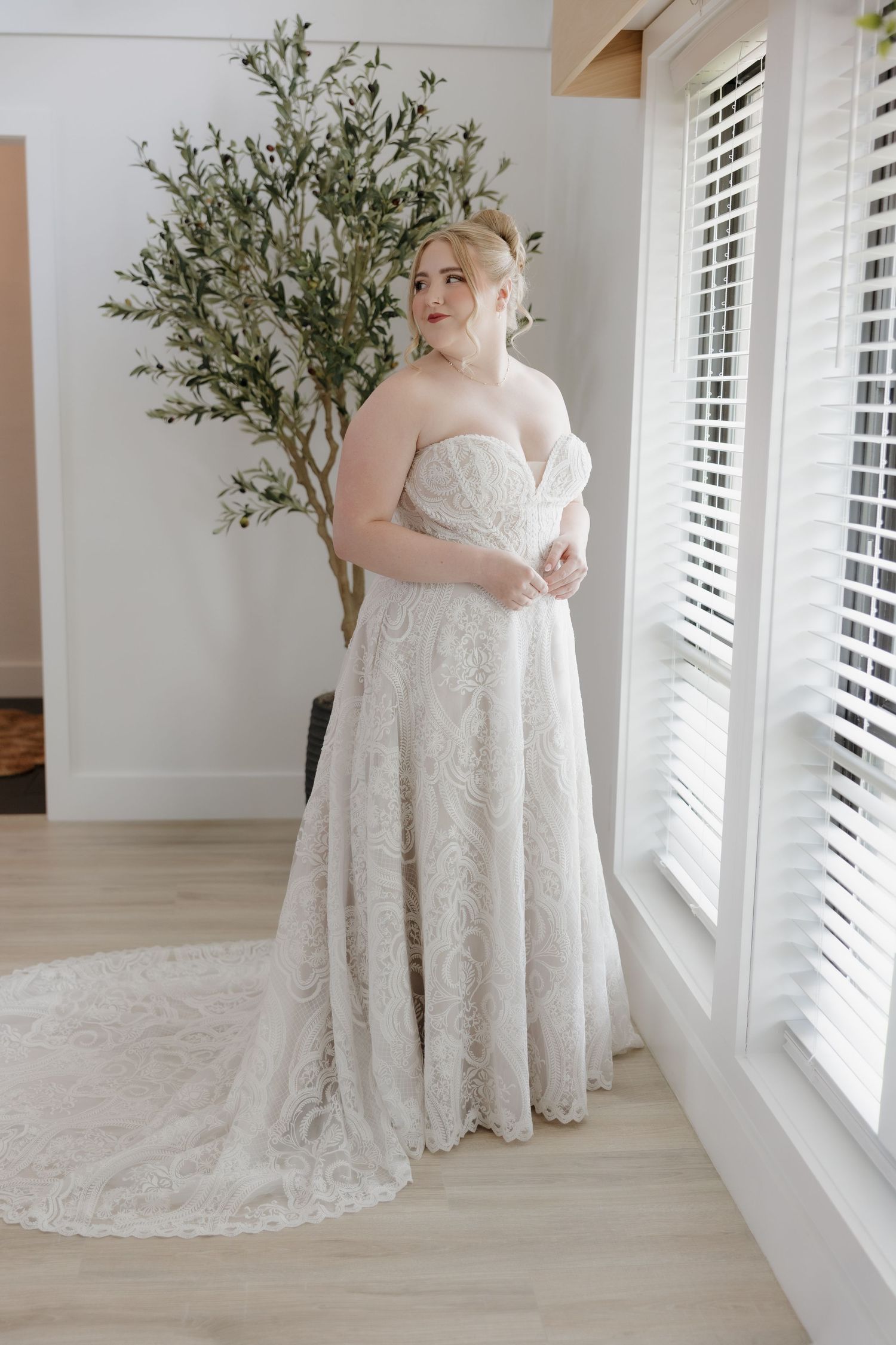 Plus-size bride wearing a strapless lace A-line wedding gown with a train, standing near white shuttered windows.