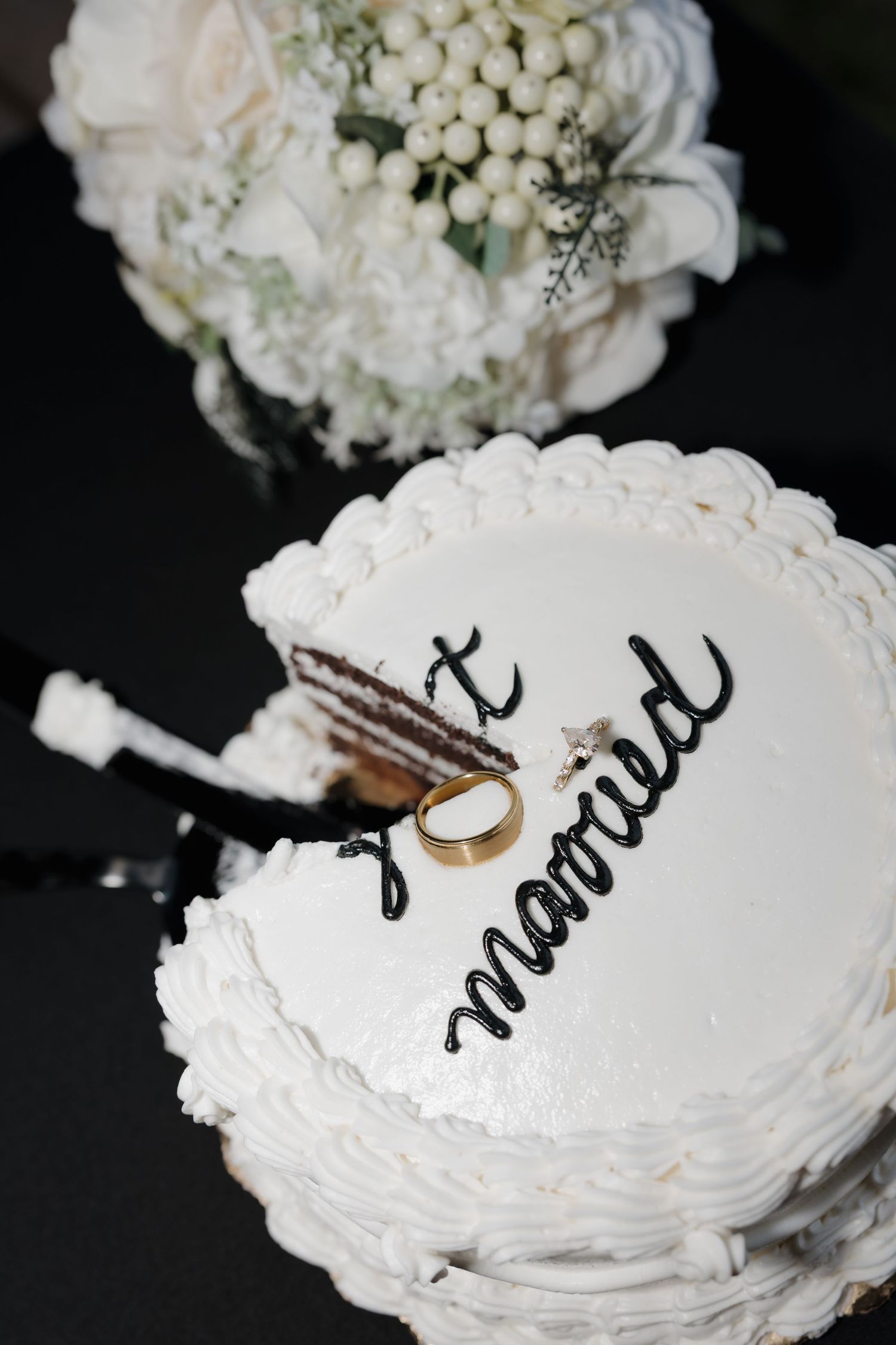 White wedding cake with 'just married' script, gold ring, and white floral bouquet on dark background.