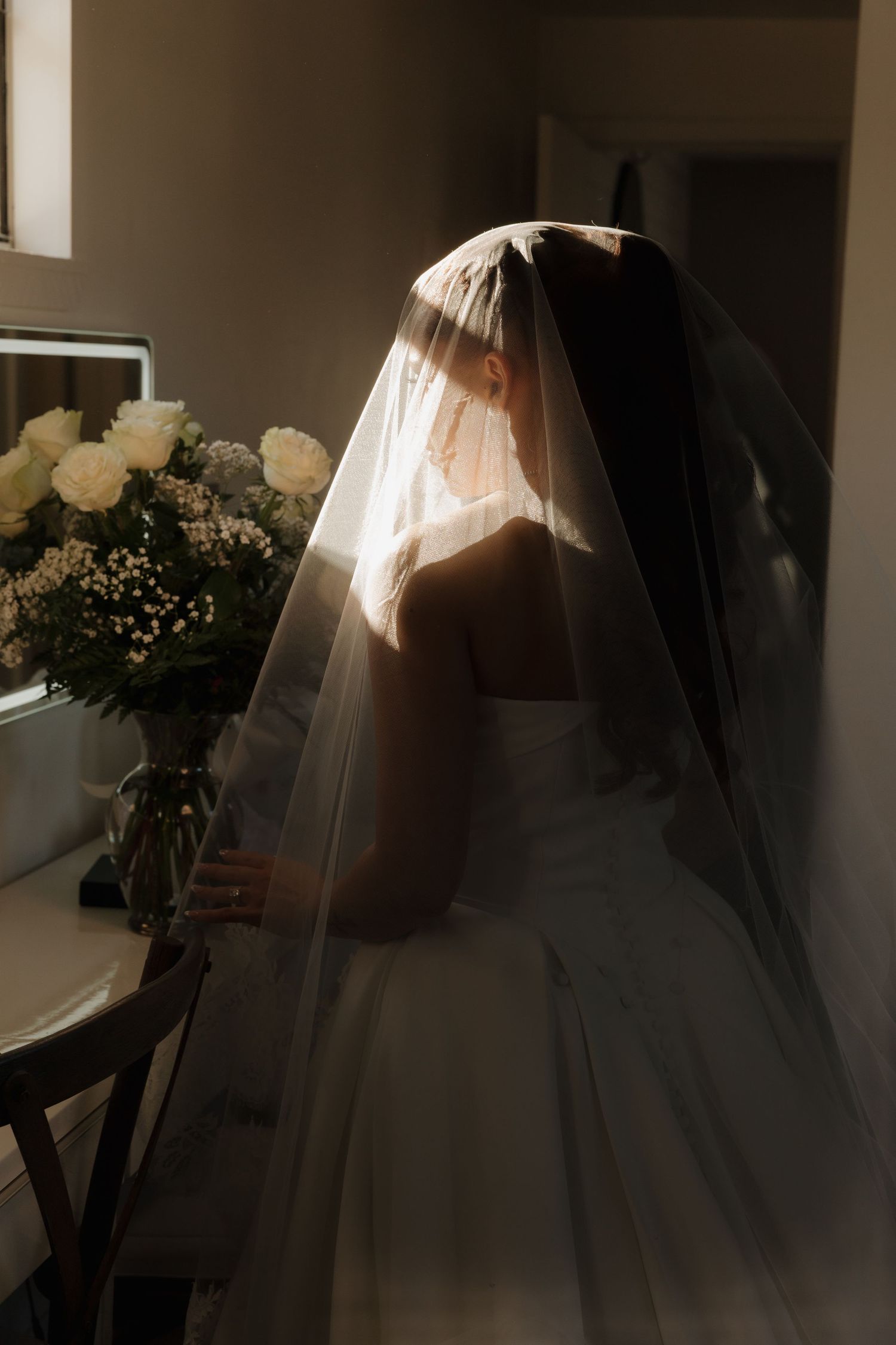 A bride in a white wedding dress and veil stands silhouetted against soft window light holding a bouquet.