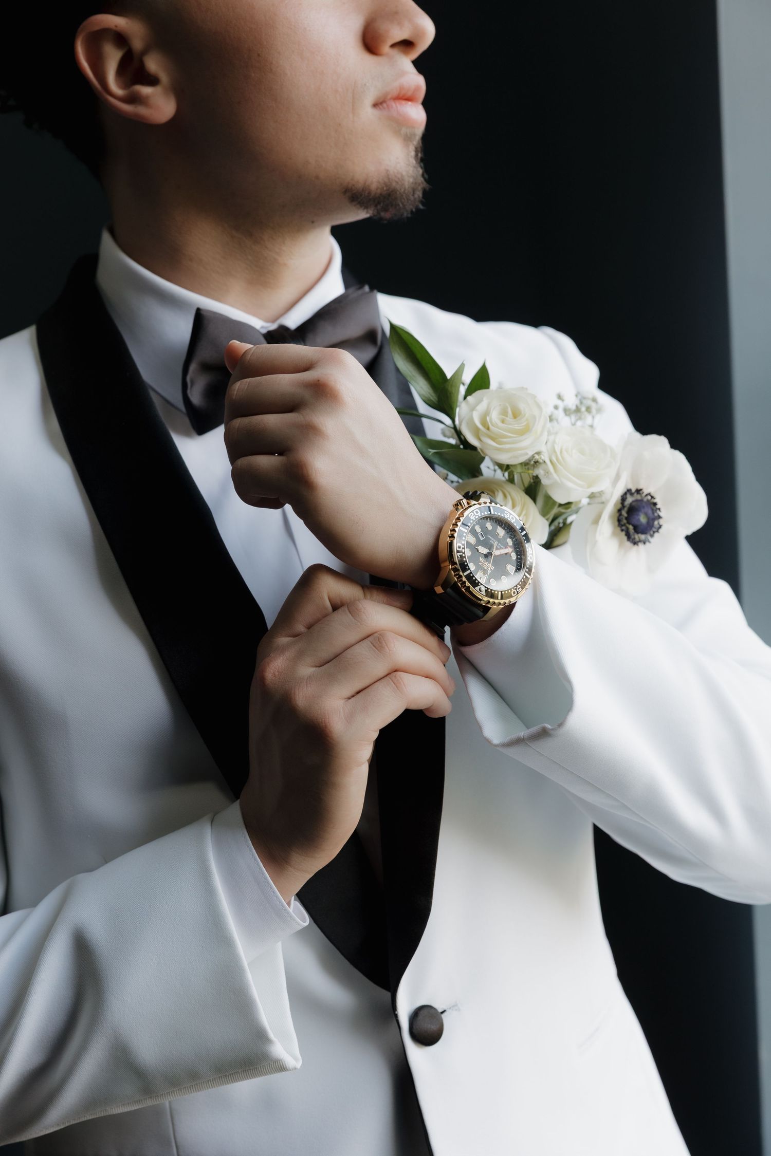 A person in a white tuxedo jacket and black bow tie adjusts their collar while wearing a wristwatch and floral boutonniere.